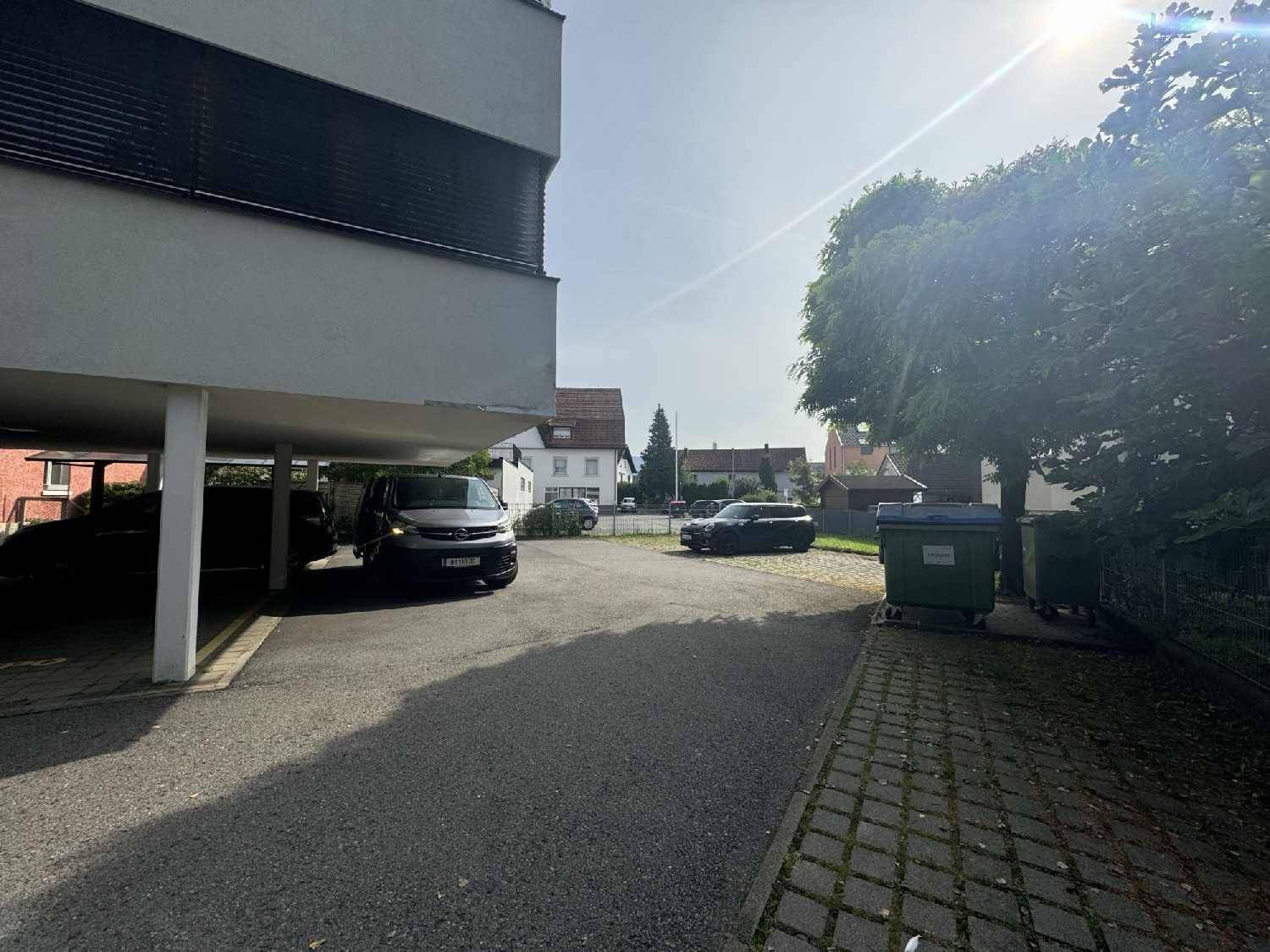Außenbereich mit überdachten Parkplätzen und einer Zufahrt, umgeben von Bäumen und weiteren Gebäuden.