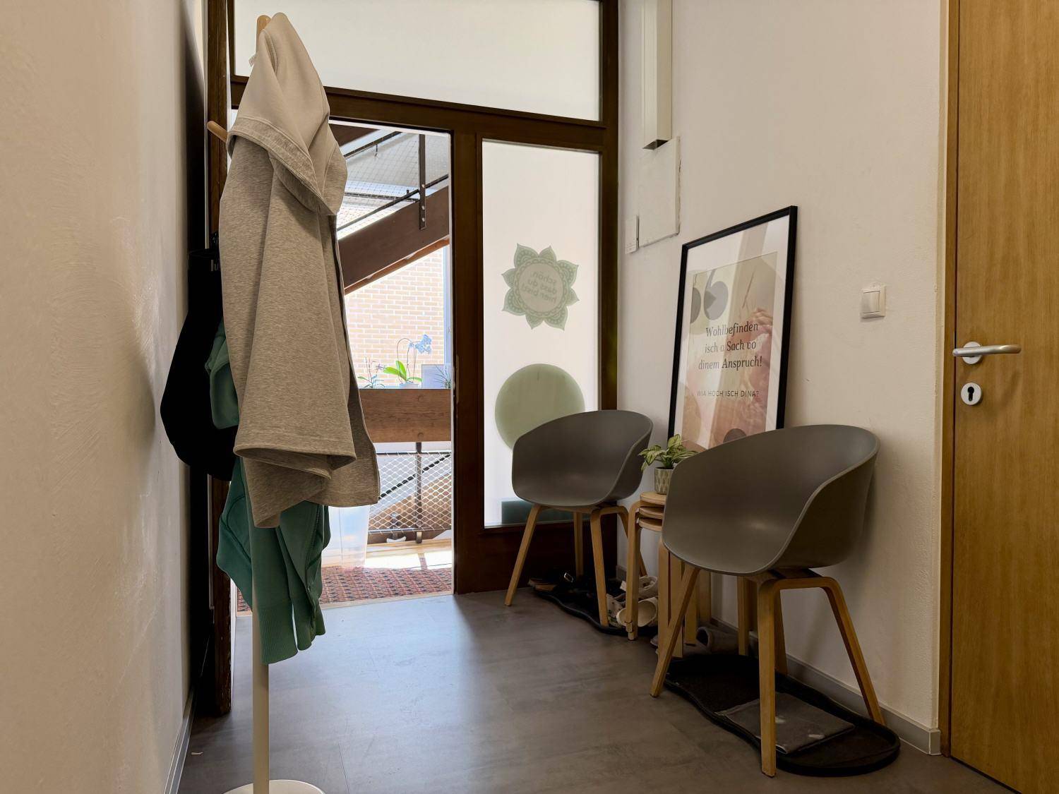 Heller Eingangsbereich mit Garderobe, modernen Sitzgelegenheiten und einer Glastür, die viel Tageslicht hereinlässt.
