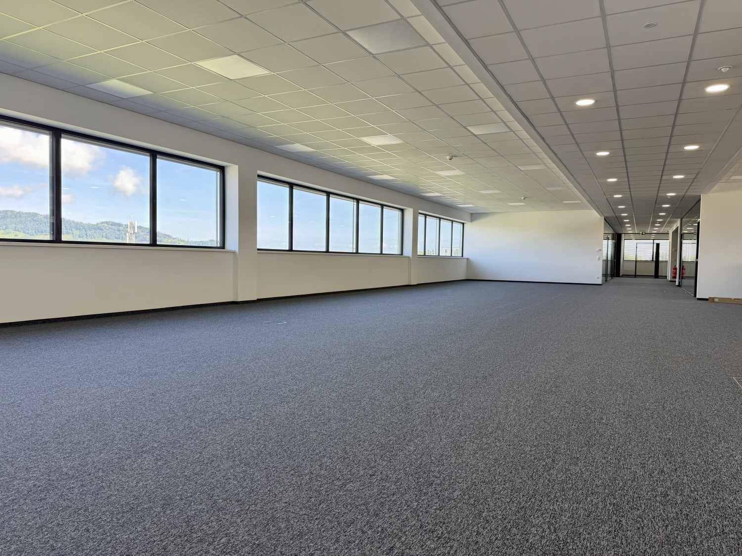 Ein weitläufiger, unmöblierter Bürobereich mit Teppichboden, vielen Fenstern und Blick auf die Landschaft.