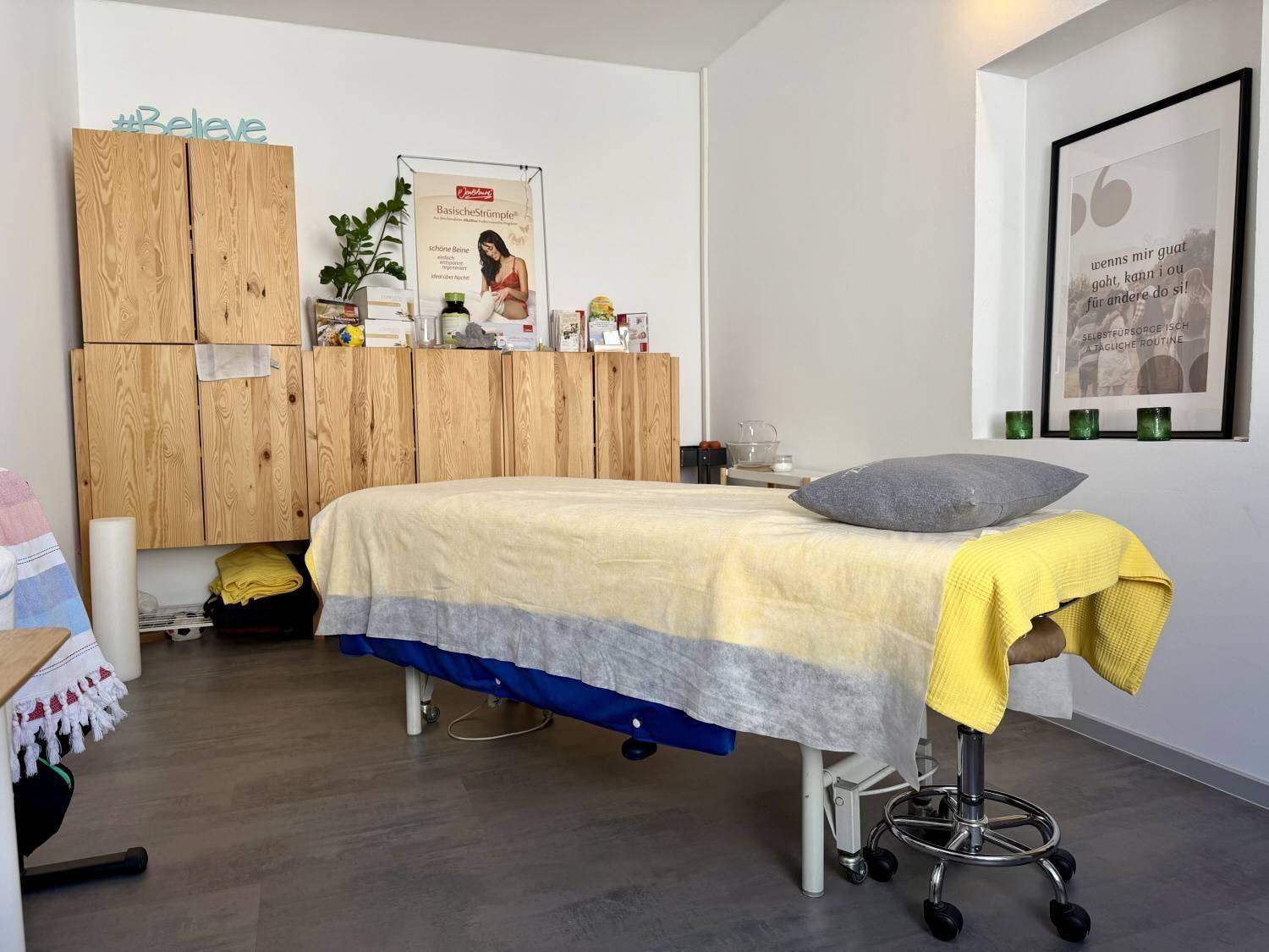 Behandlungsraum mit einer Massageliege, rustikalen Holzschränken und einem gerahmten Bild an der weißen Wand.