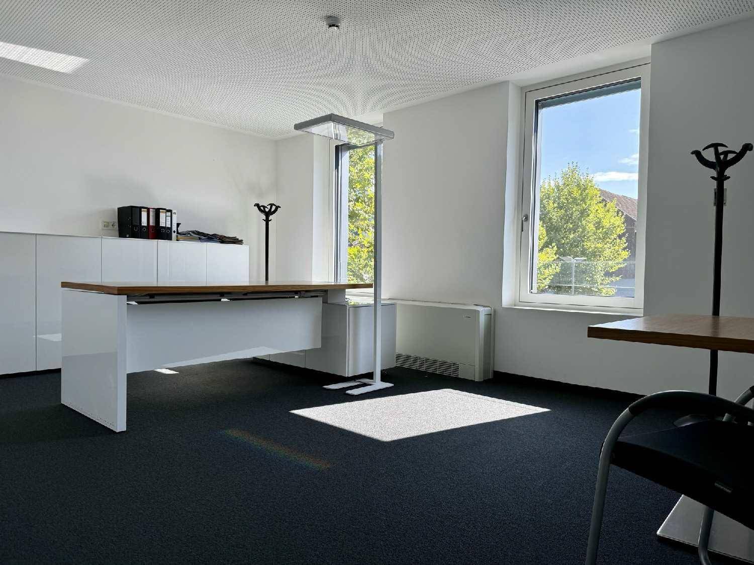 Helles Büro mit Schreibtisch, Schrank und zwei großen Fenstern, die viel Tageslicht hereinlassen.