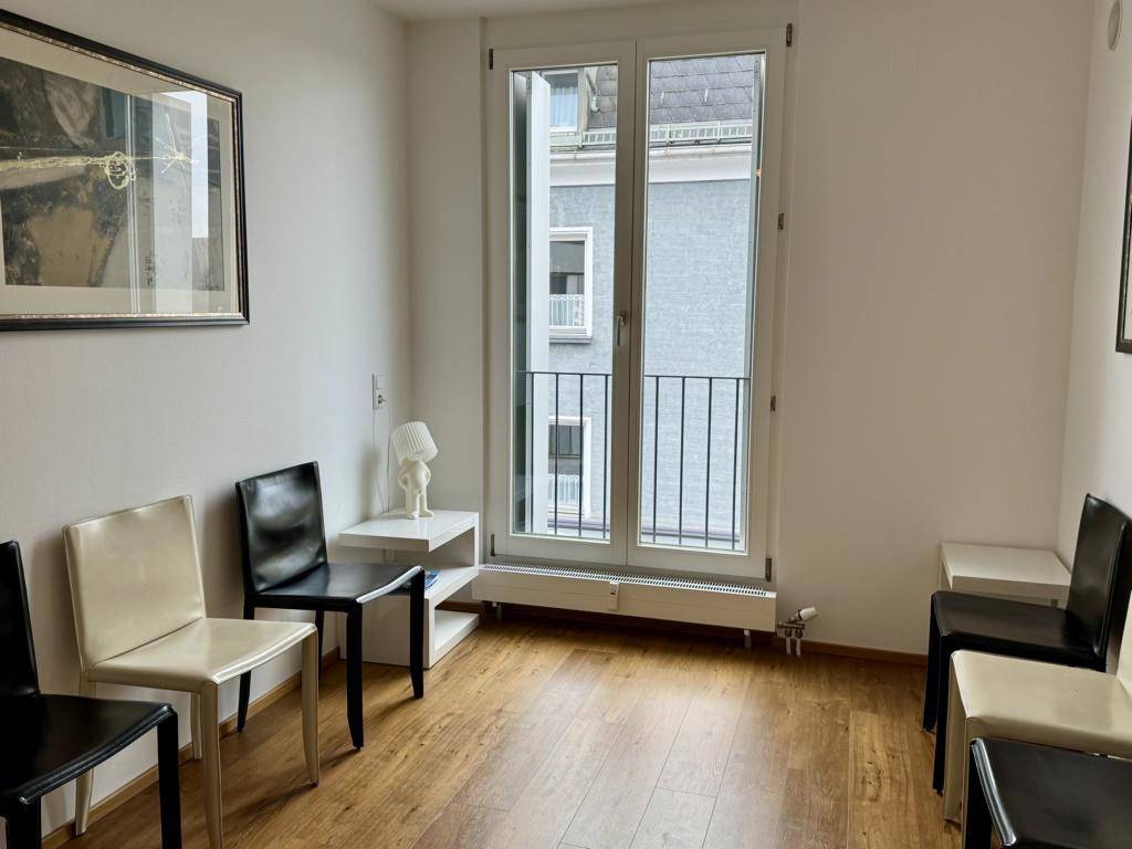 Wartezimmer mit Holzboden, mehreren Stühlen und einem französischen Balkon, der viel Tageslicht hereinlässt.
