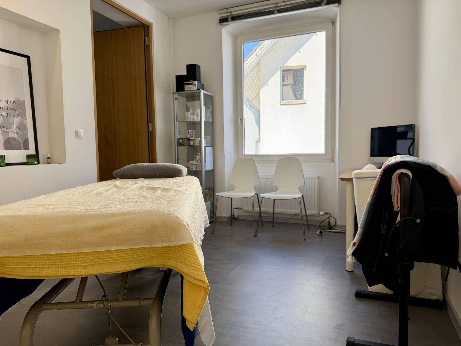 Heller Behandlungsraum mit Massageliege, großem Fenster und zwei Stühlen, der eine ruhige und einladende Atmosphäre bietet.