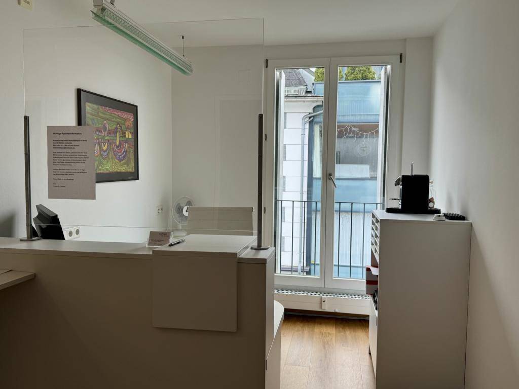 Empfangsbereich mit Schreibtisch, Schutzscheibe und einem Fenster, das viel Tageslicht hereinlässt und einen Balkon zeigt.