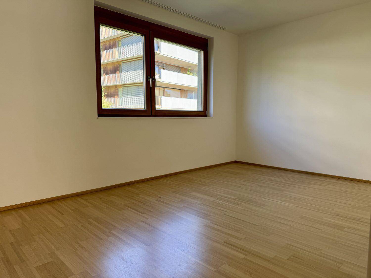 Leeres Schlafzimmer mit Holzboden und einem Fenster, das den Blick auf ein anderes Gebäude freigibt.