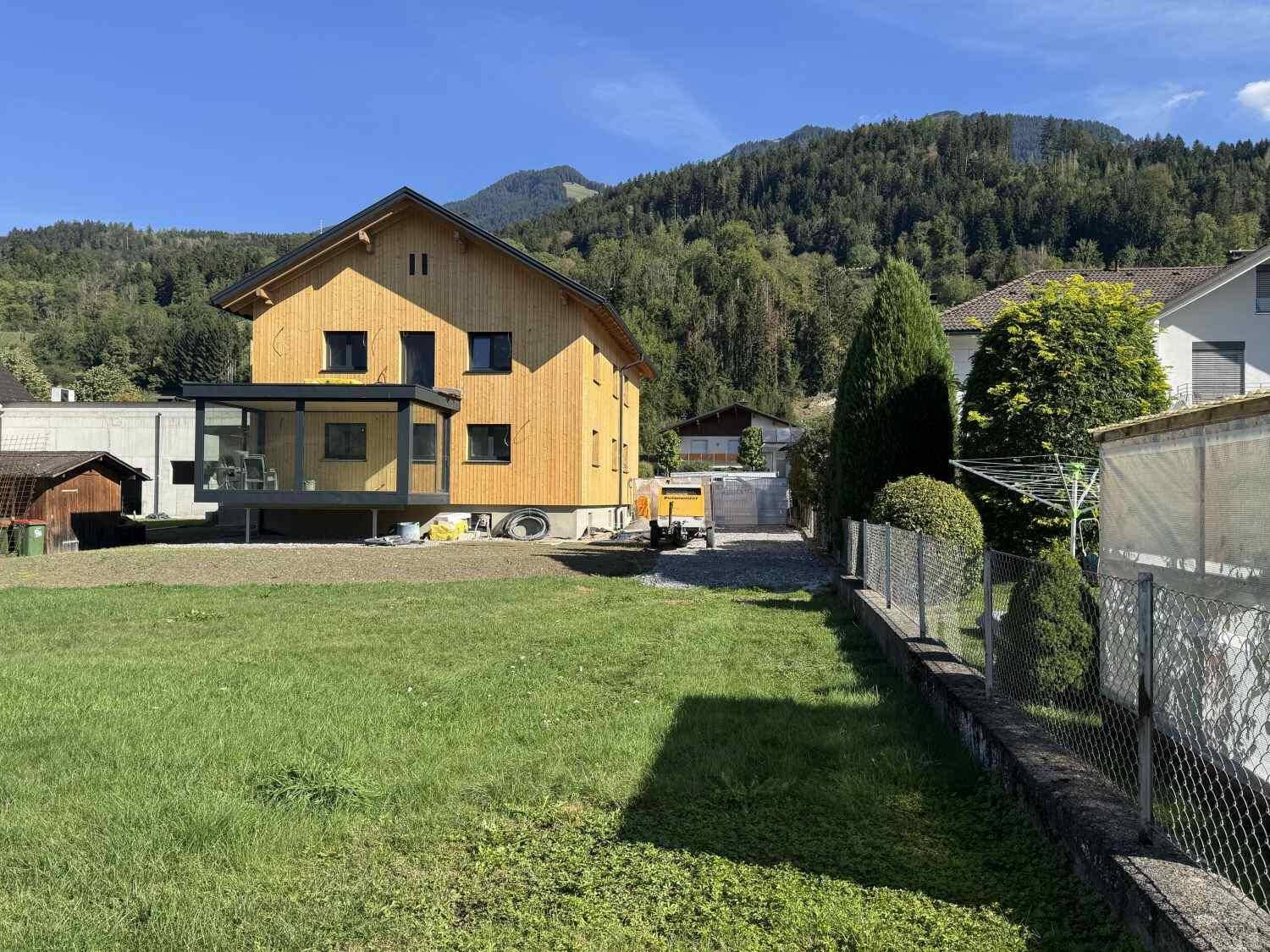 Neues Holzhaus mit moderner Glaserweiterung und Rasenfläche vor bewaldeten Bergen.