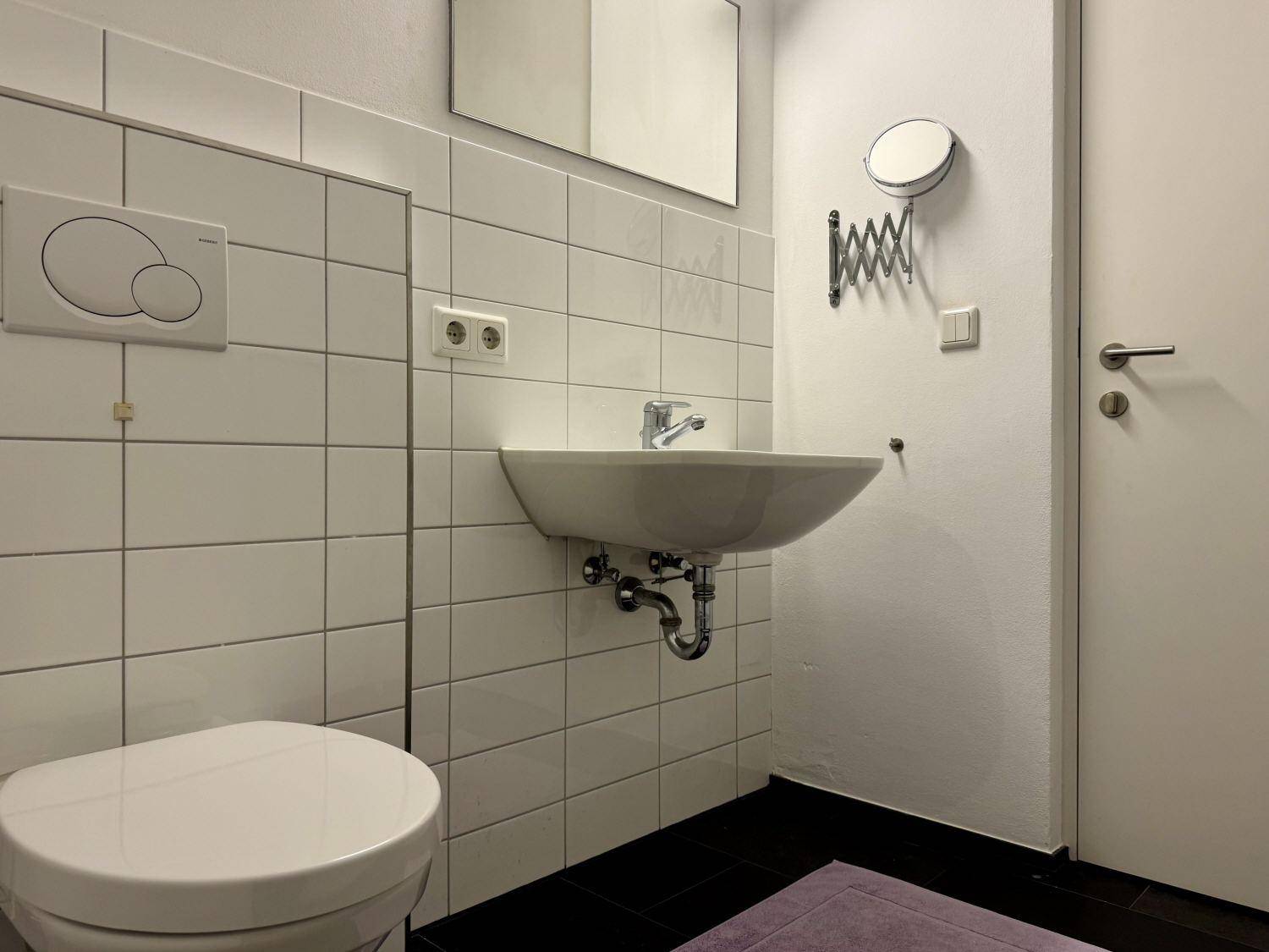 Badezimmer mit Waschbecken, Wand-WC und weißen Fliesen, ausgestattet mit einem Kosmetikspiegel.