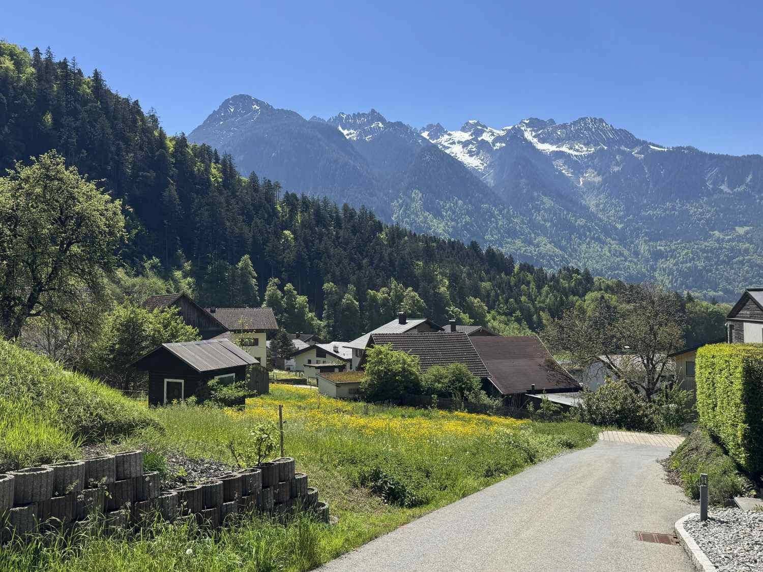 Ländliche Straße führt durch eine Siedlung mit Häusern und grünen Wiesen, umgeben von Bergen.