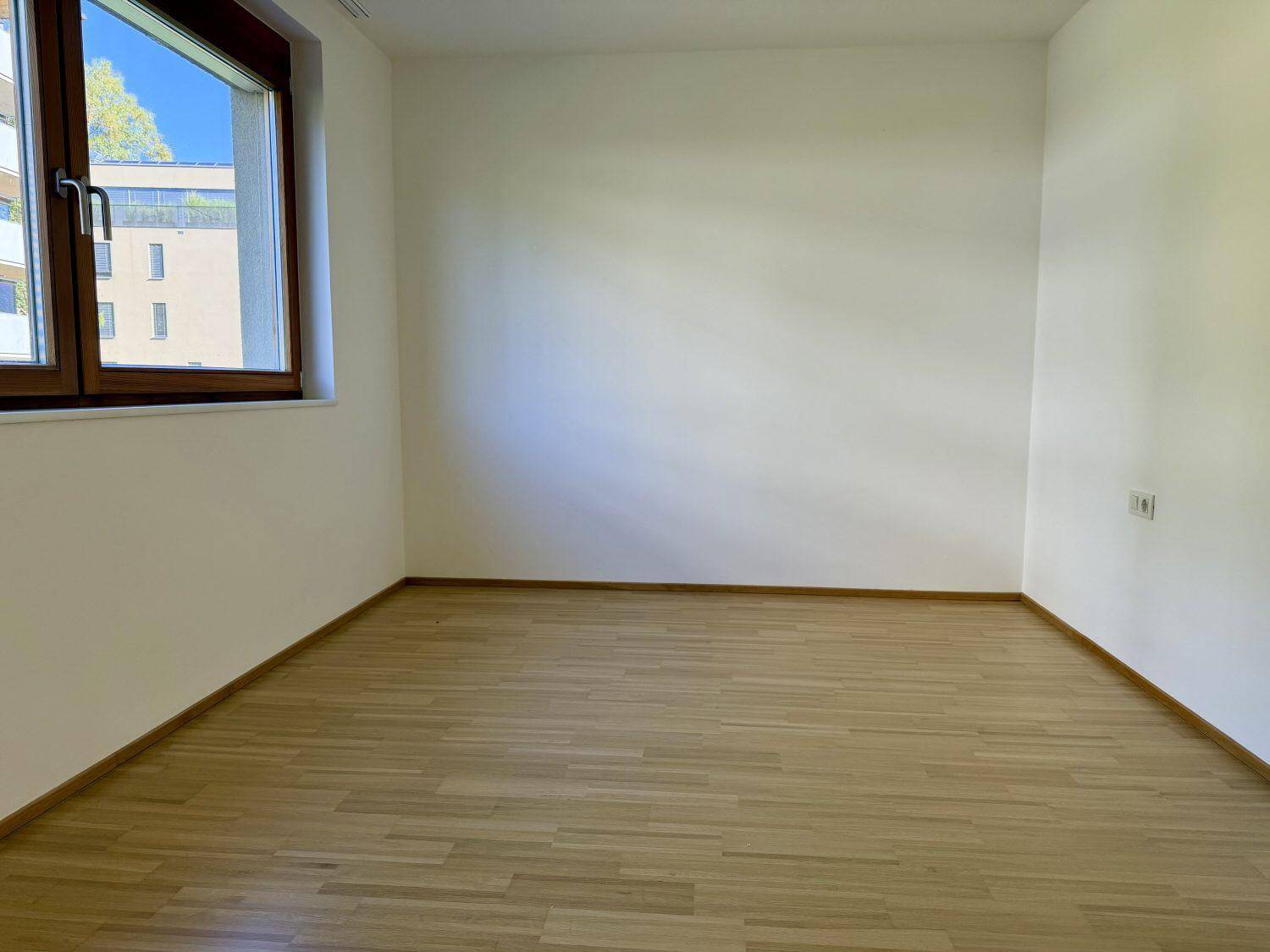 Helles, leeres Zimmer mit Holzboden und einem Fenster, das viel Tageslicht hereinlässt.