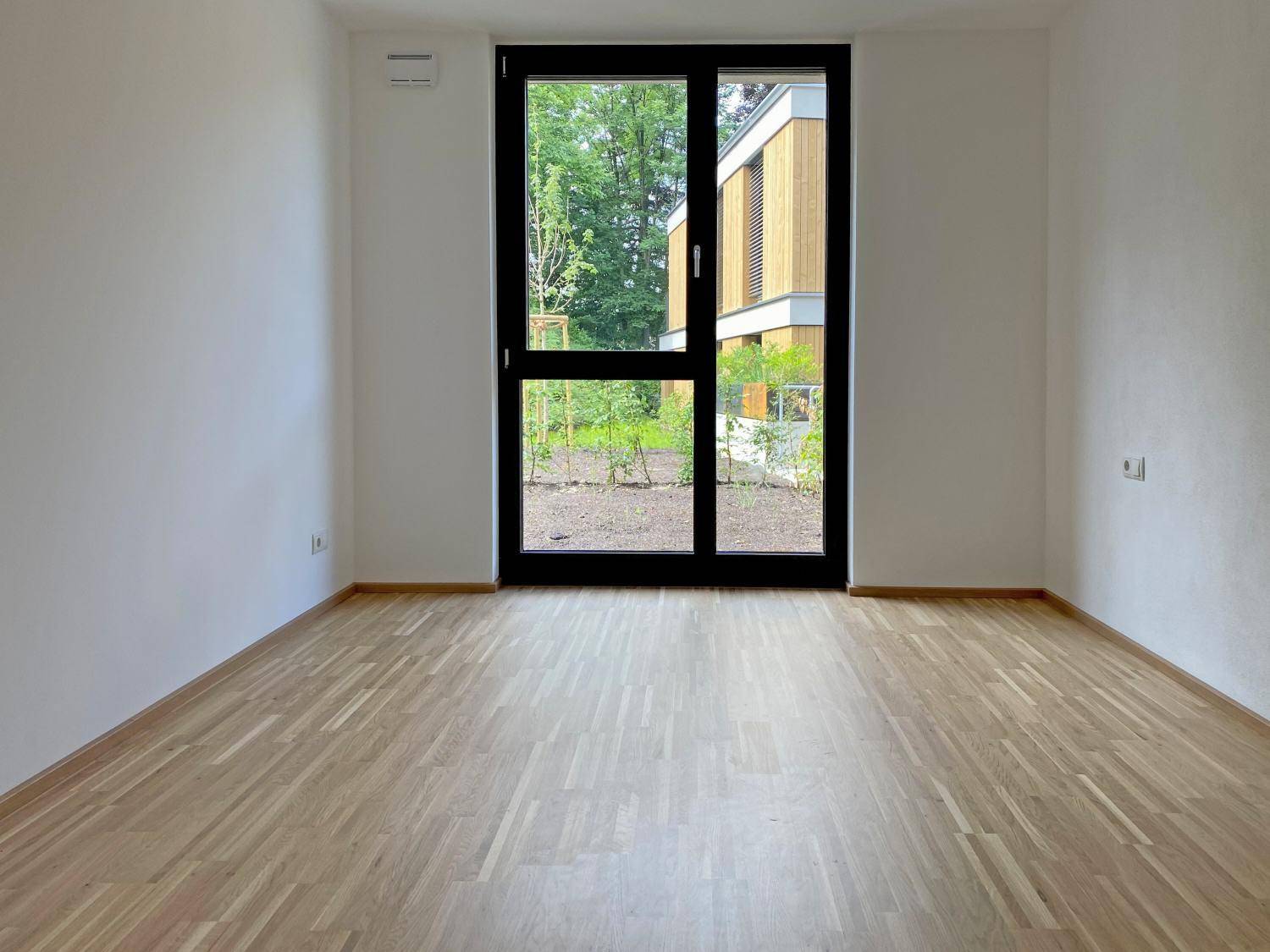 Helles Zimmer mit Parkettboden und großem Fenster mit Blick ins Grüne.