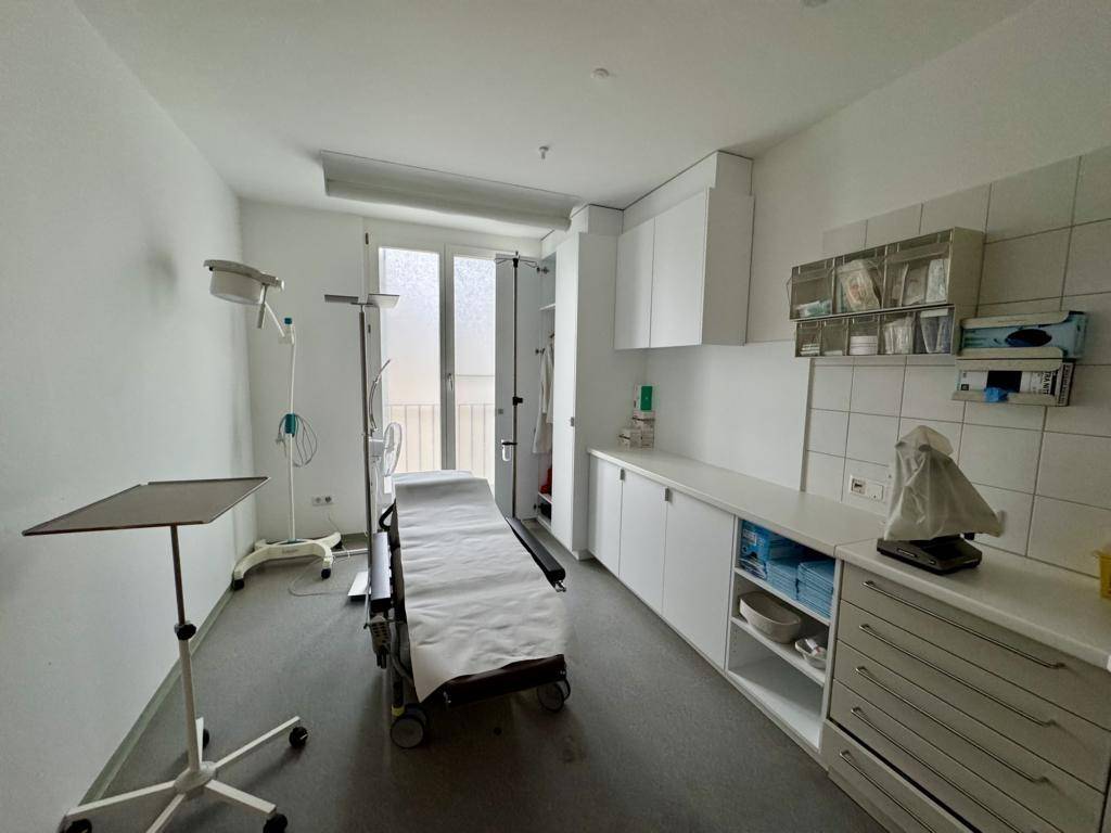 Behandlungszimmer mit medizinischer Liege, weißen Schränken und Fliesen an der Wand, funktional und sauber.