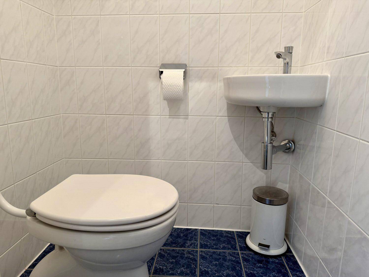 Separates WC mit weißen Wandfliesen, kleinem Waschbecken und blauen Bodenfliesen.