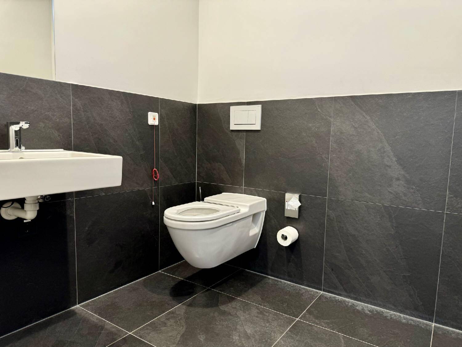 Modernes WC mit dunkelgrauen Fliesen, wandhängender Toilette und kleinem Waschbecken, inklusive Notrufsystem.