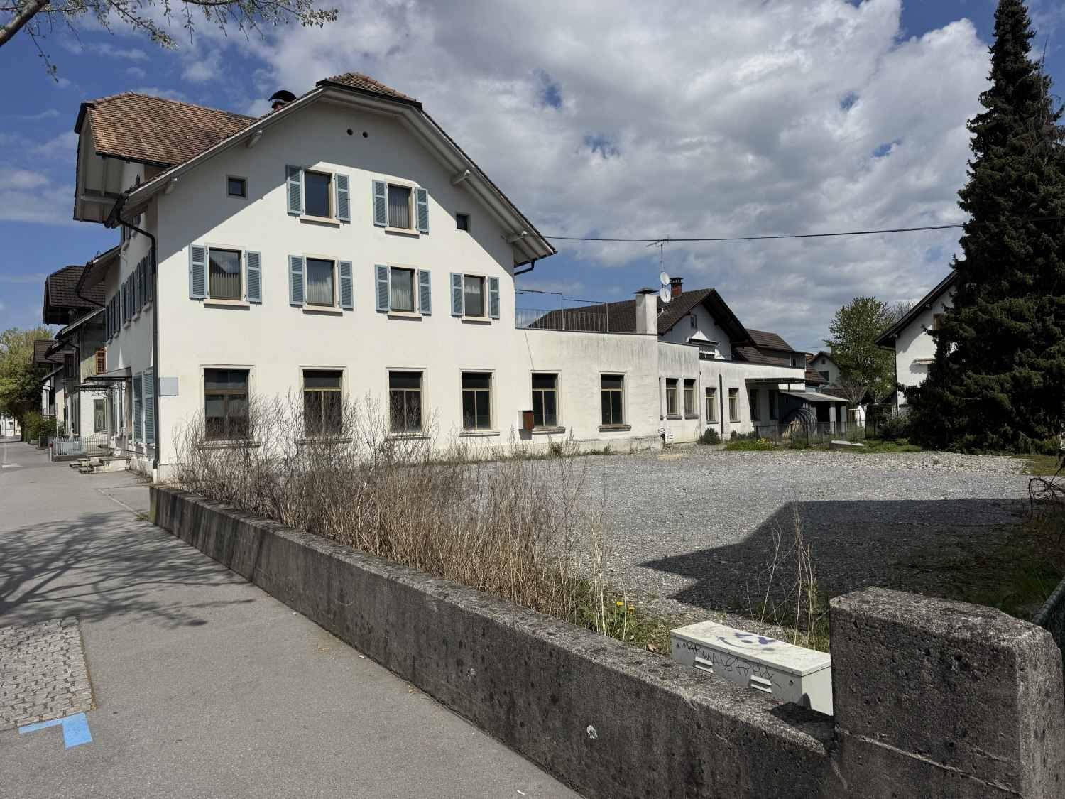 Rückansicht des Gebäudes mit einem großen Schotterplatz, umgeben von niedriger Vegetation.