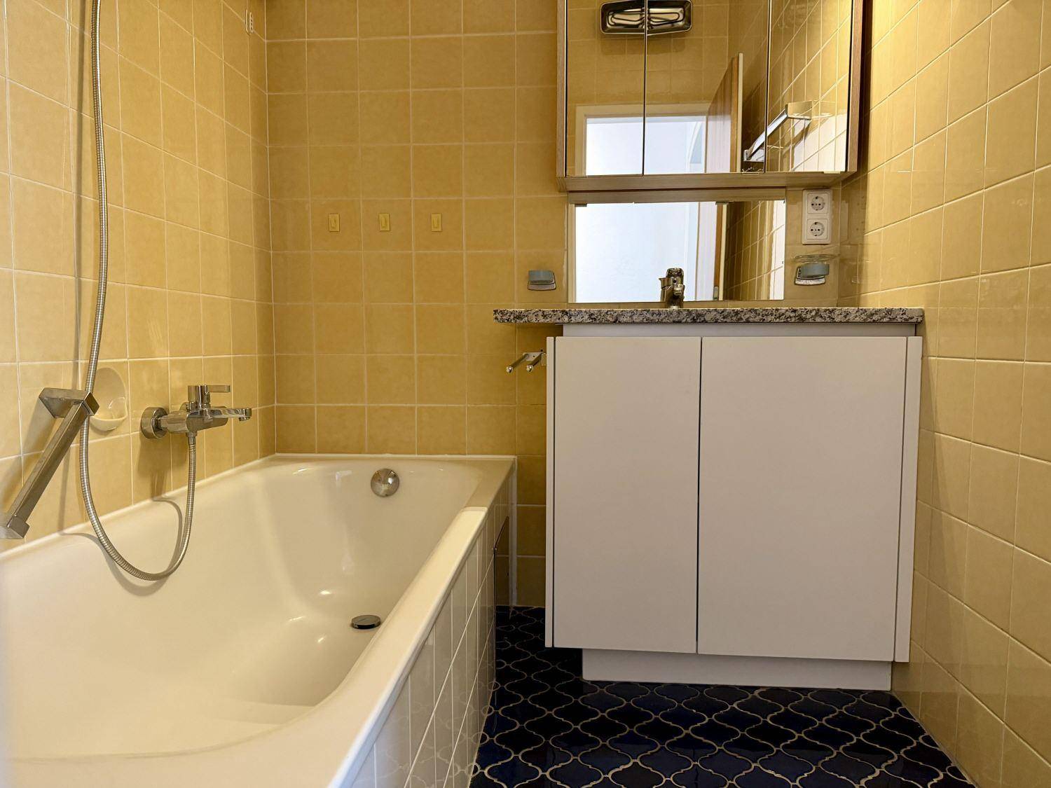 Badezimmer mit gelben Wandfliesen, blauen Bodenfliesen, Badewanne und Waschtisch.
