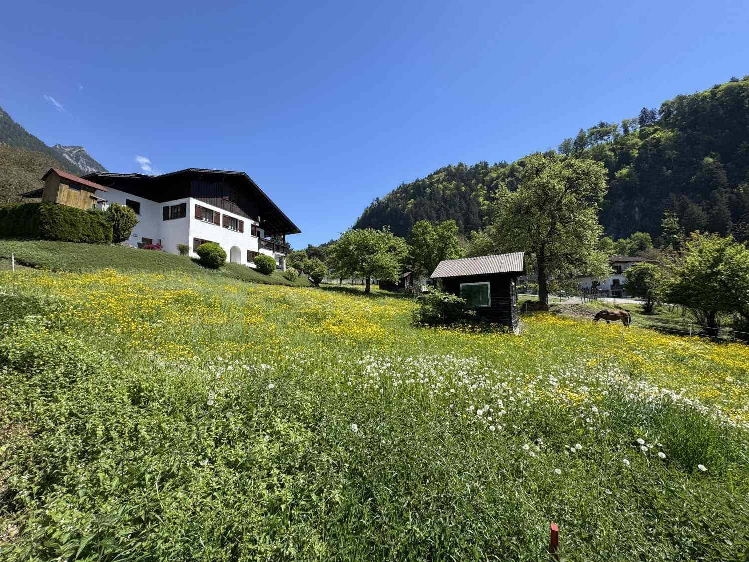 Traditionelles Haus mit Balkon und weiten grünen Wiesen vor einer beeindruckenden Berglandschaft.