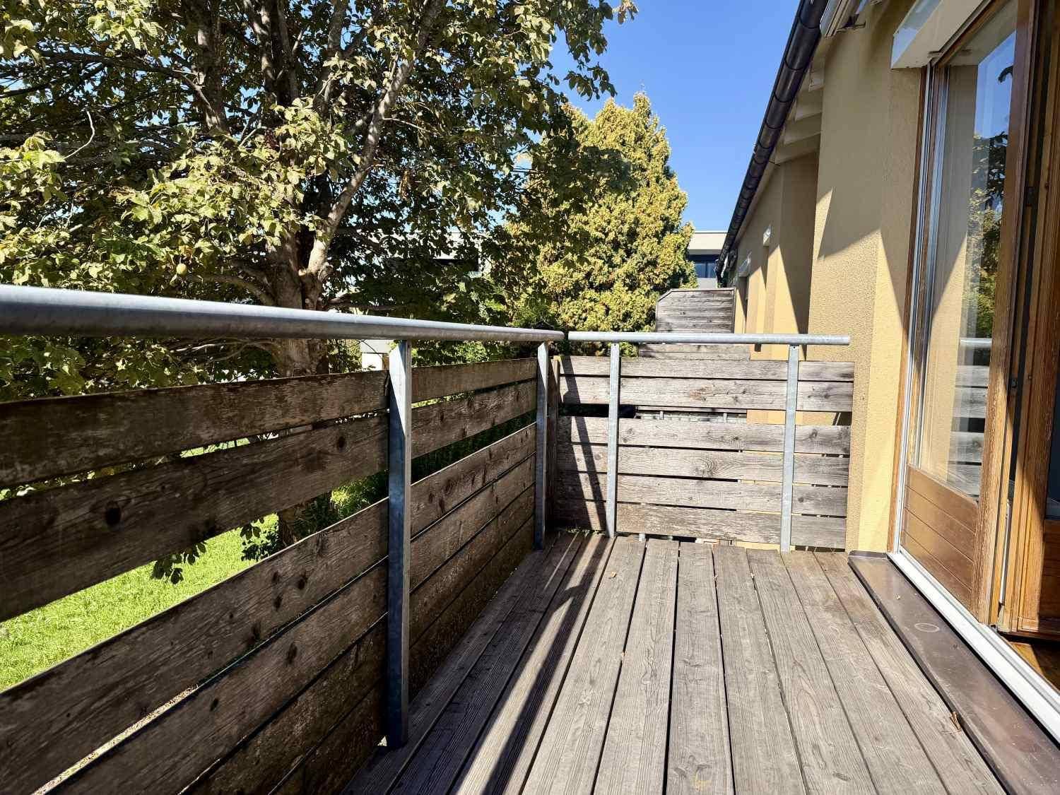 Sonniger Balkon mit Holzboden und Holzgeländer, umgeben von üppigem Grün und Bäumen.