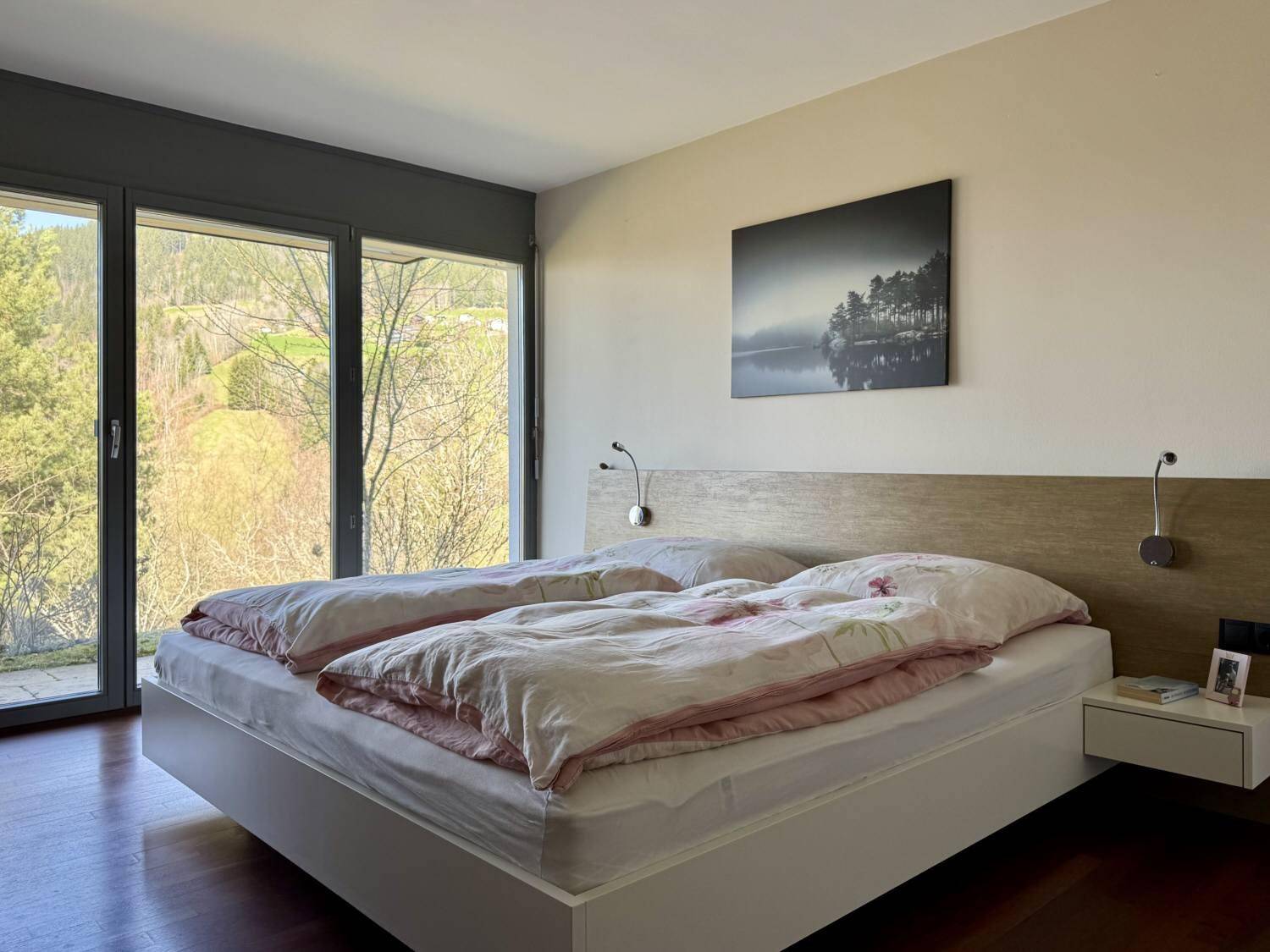 Helles Schlafzimmer mit Doppelbett, Holzboden und großen Fenstern mit Blick ins Grüne.