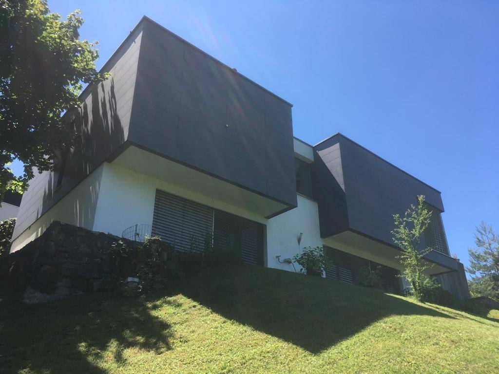 Moderne Architektur des Hauses mit dunkler Fassadenverkleidung und großen Fenstern auf einem grünen Hang.