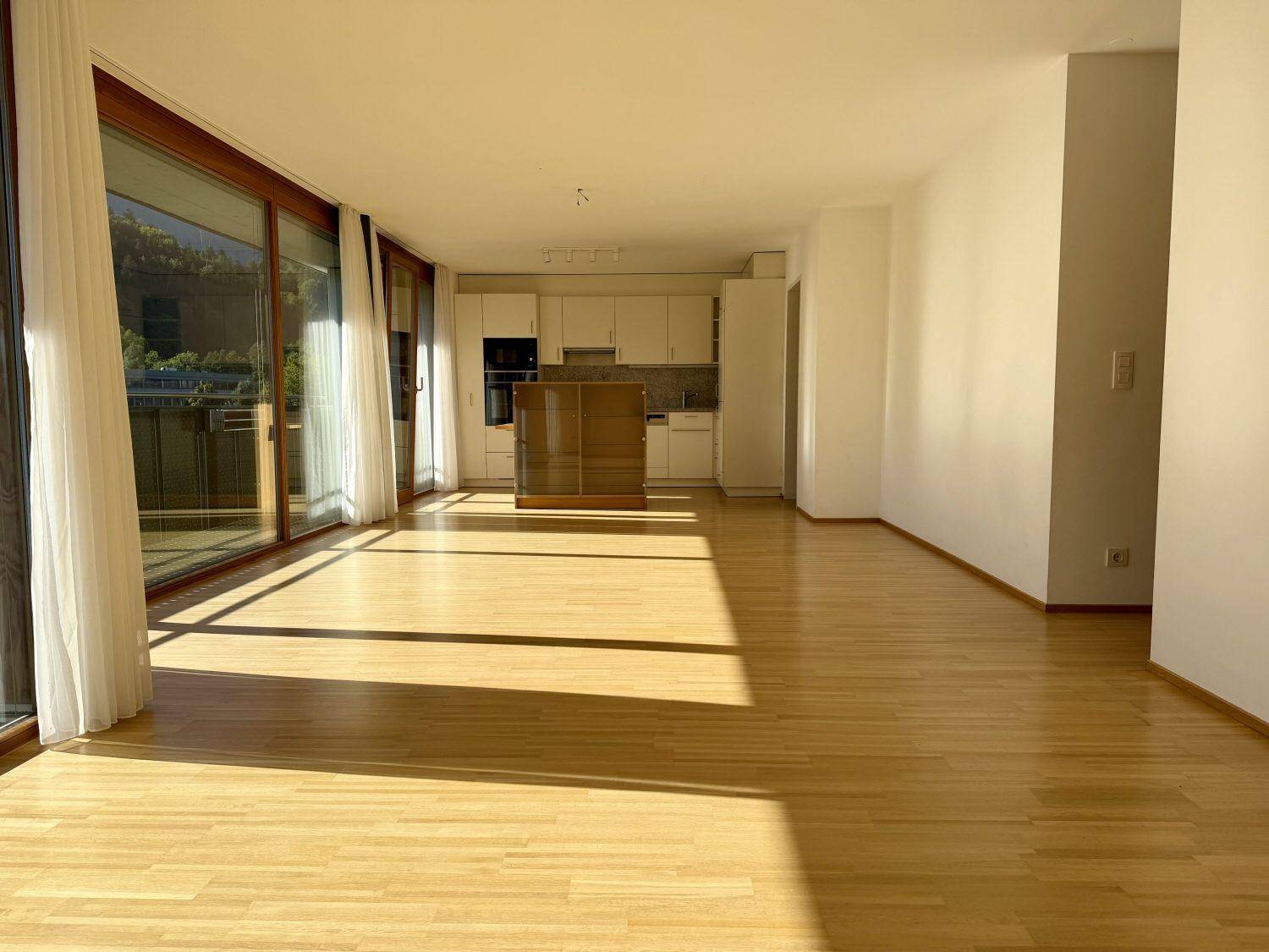 Offener Wohnbereich mit Holzboden und integrierter Küche im Hintergrund, durchflutet von Sonnenlicht.