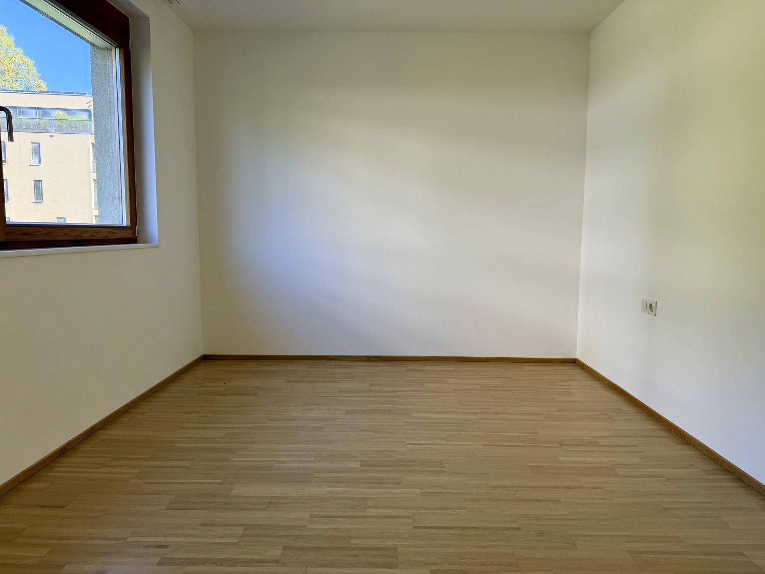 Helles, leeres Zimmer mit Holzboden und einem Fenster, das viel Tageslicht hereinlässt.
