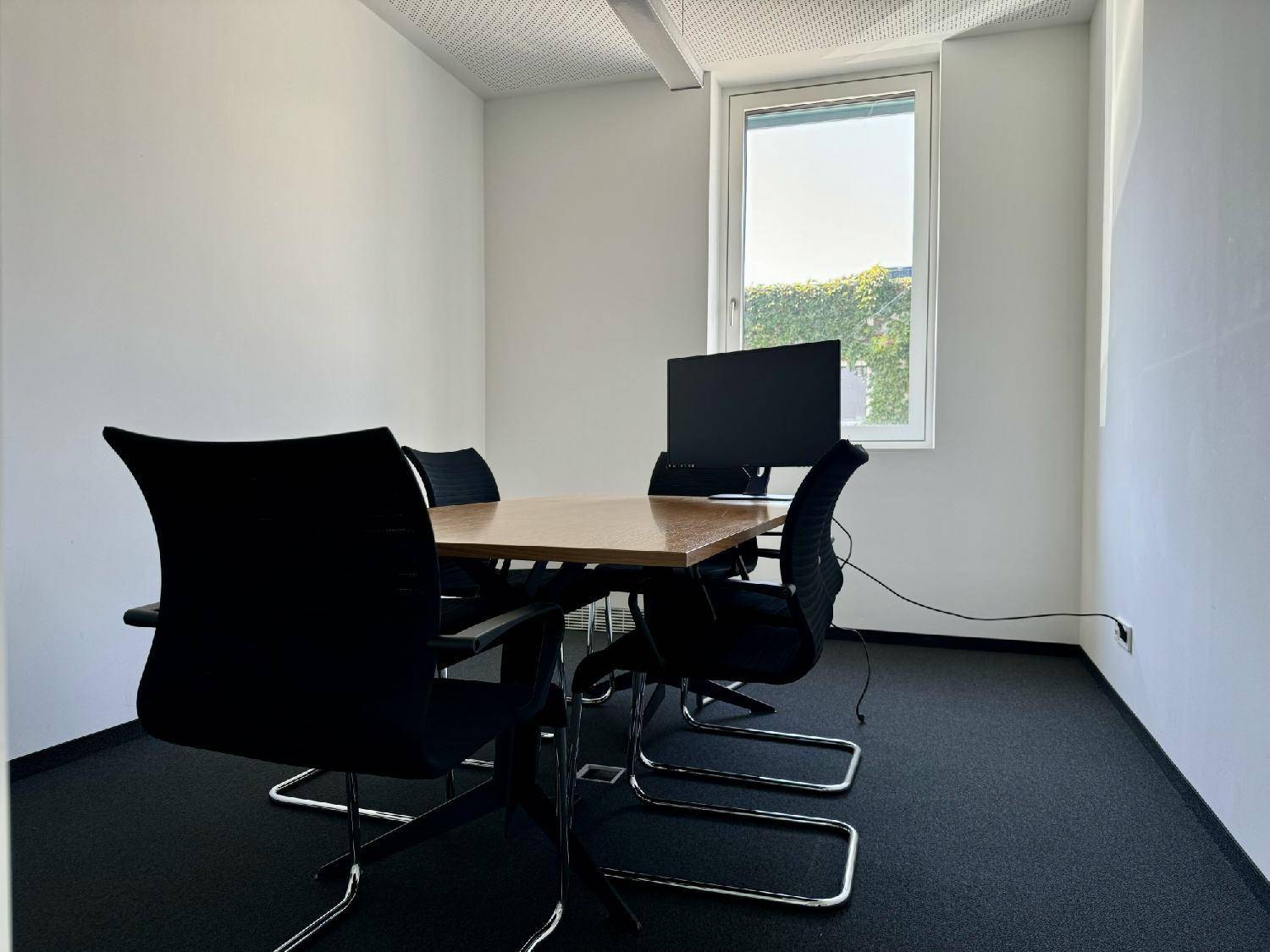 Besprechungsraum mit Konferenztisch, Stühlen, Monitor und Teppichboden, hell durch das Fenster.
