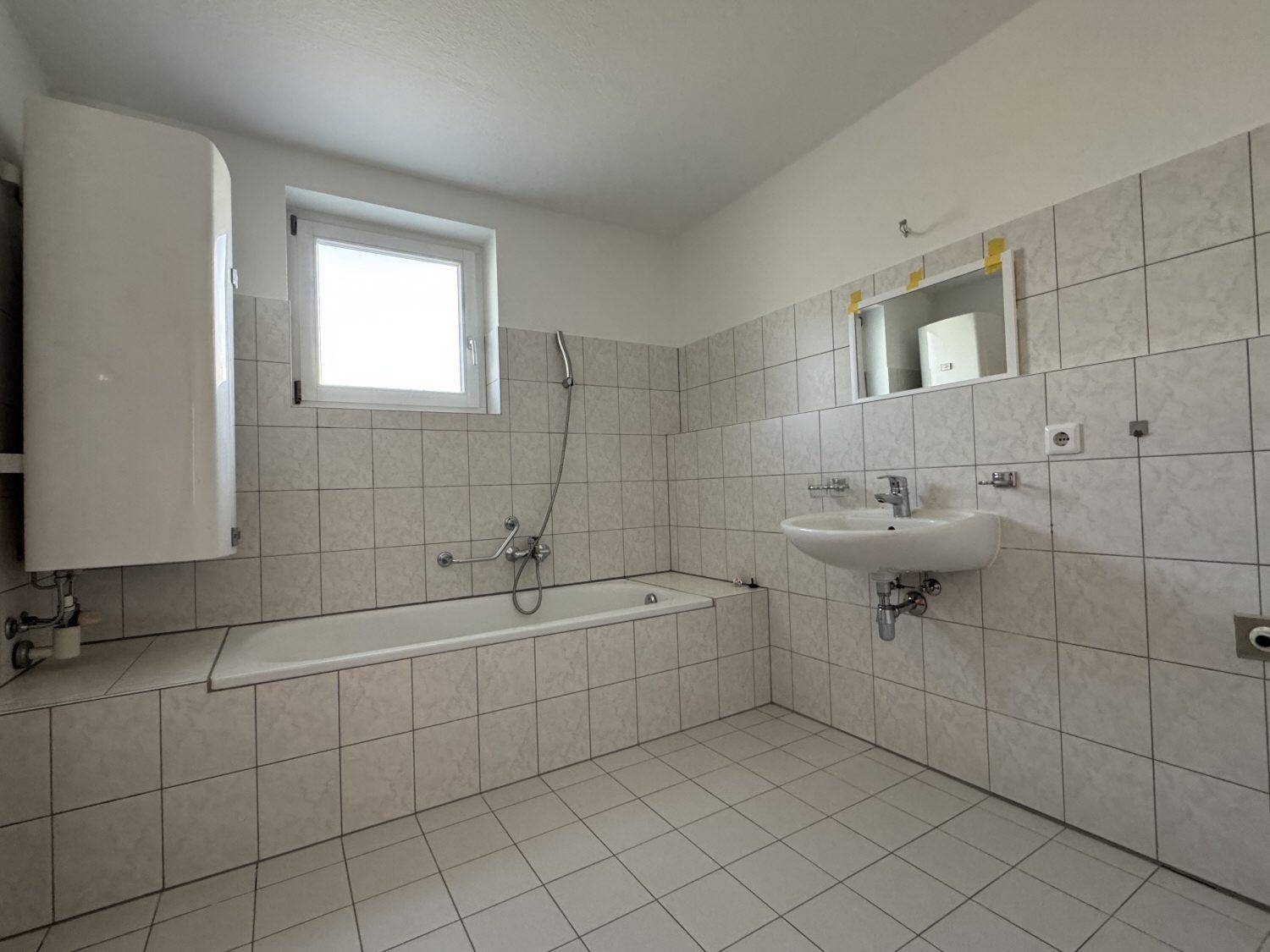 Gefliestes Badezimmer mit Badewanne, Waschbecken, Fenster und Warmwasserboiler.