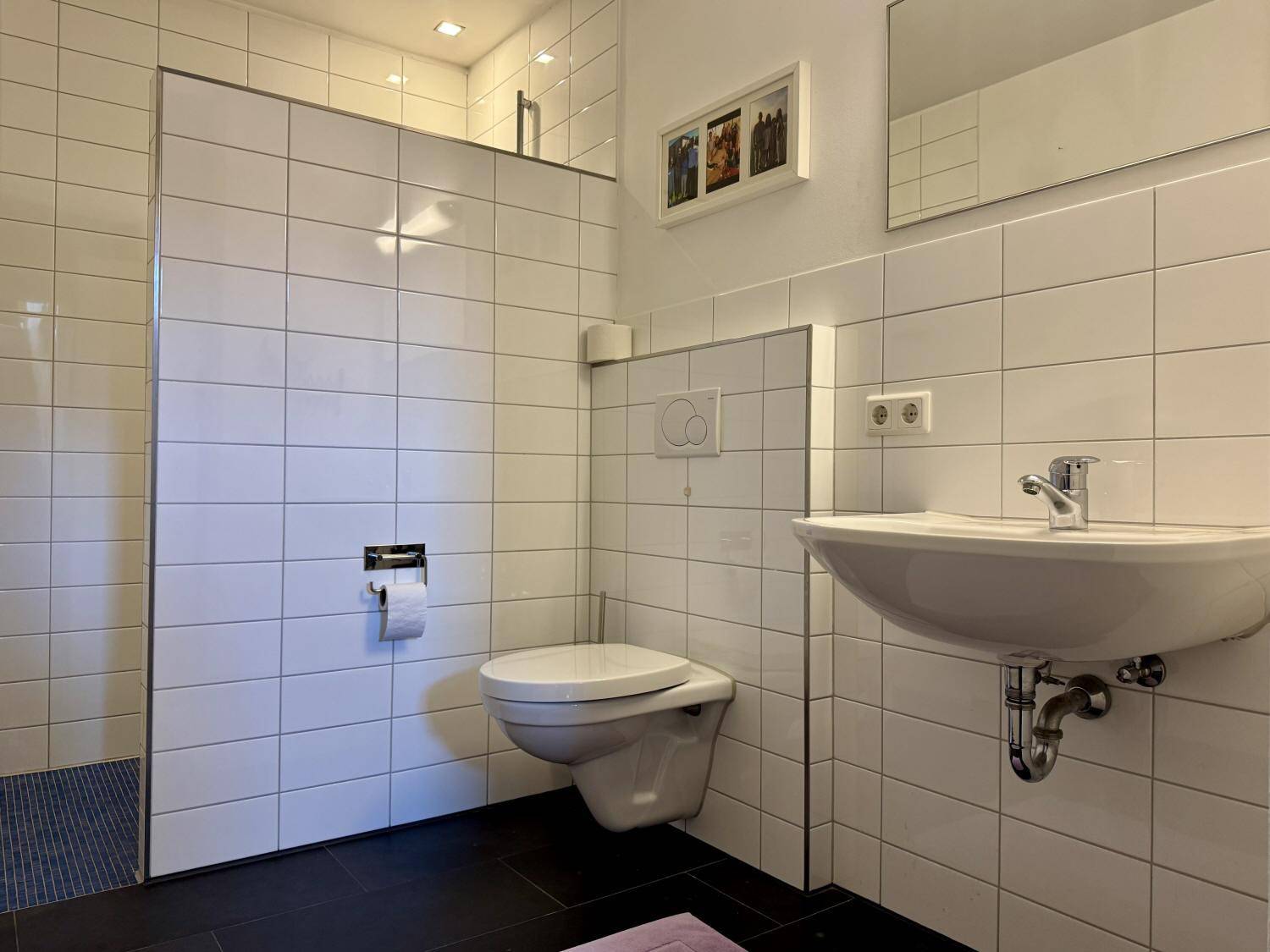 Helles Badezimmer mit Wand-WC, Waschbecken und weißen Wandfliesen.