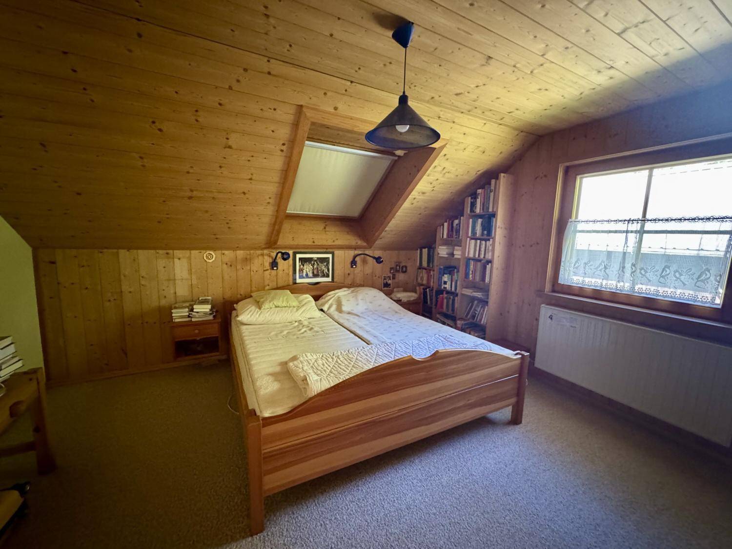 Gemütliches Schlafzimmer im Dachgeschoss mit Holzdachschrägen, Teppichboden und Bücherregal.