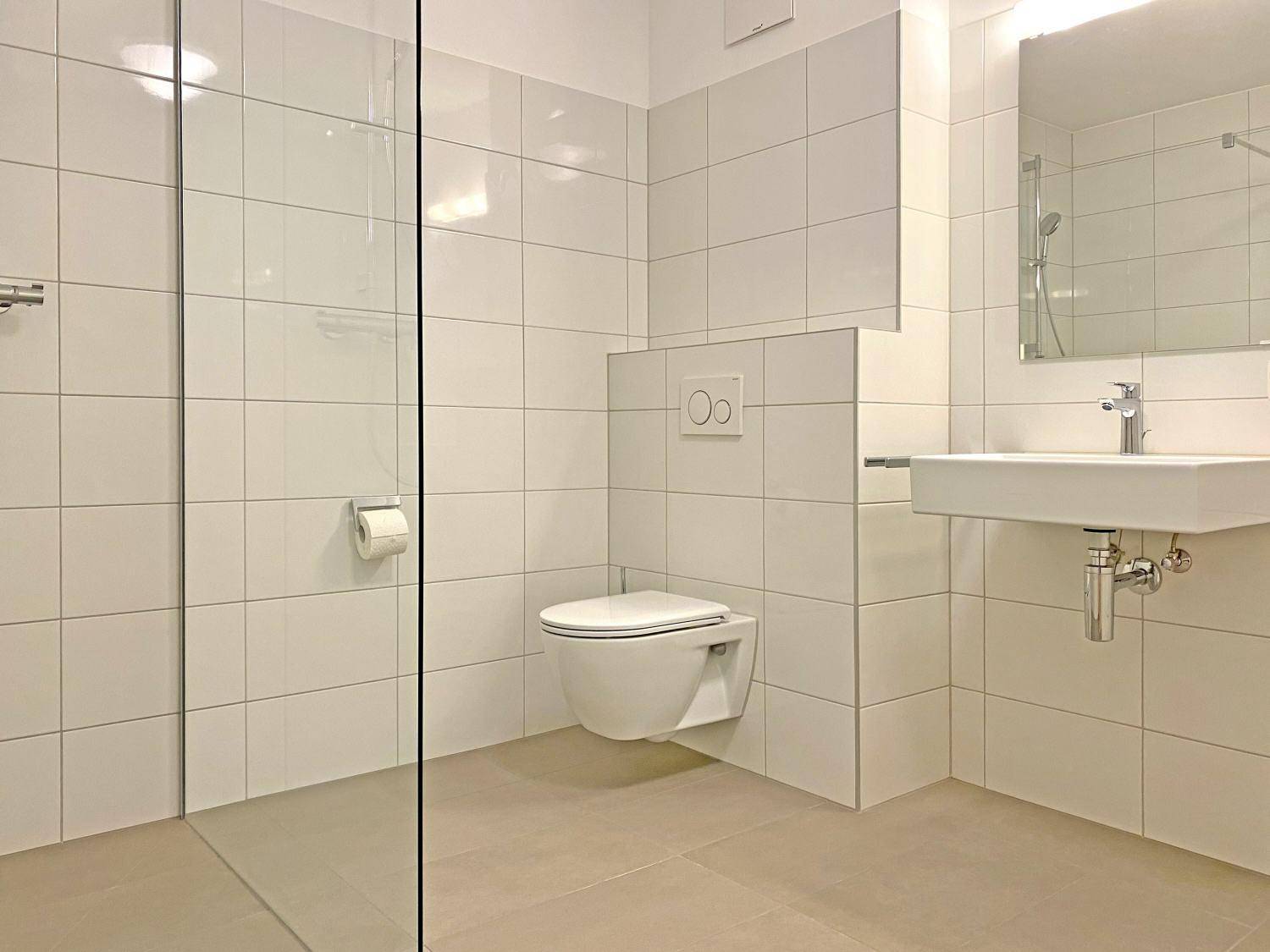 Modernes Badezimmer mit weißen Fliesen, wandhängendem WC und ebenerdiger Dusche.