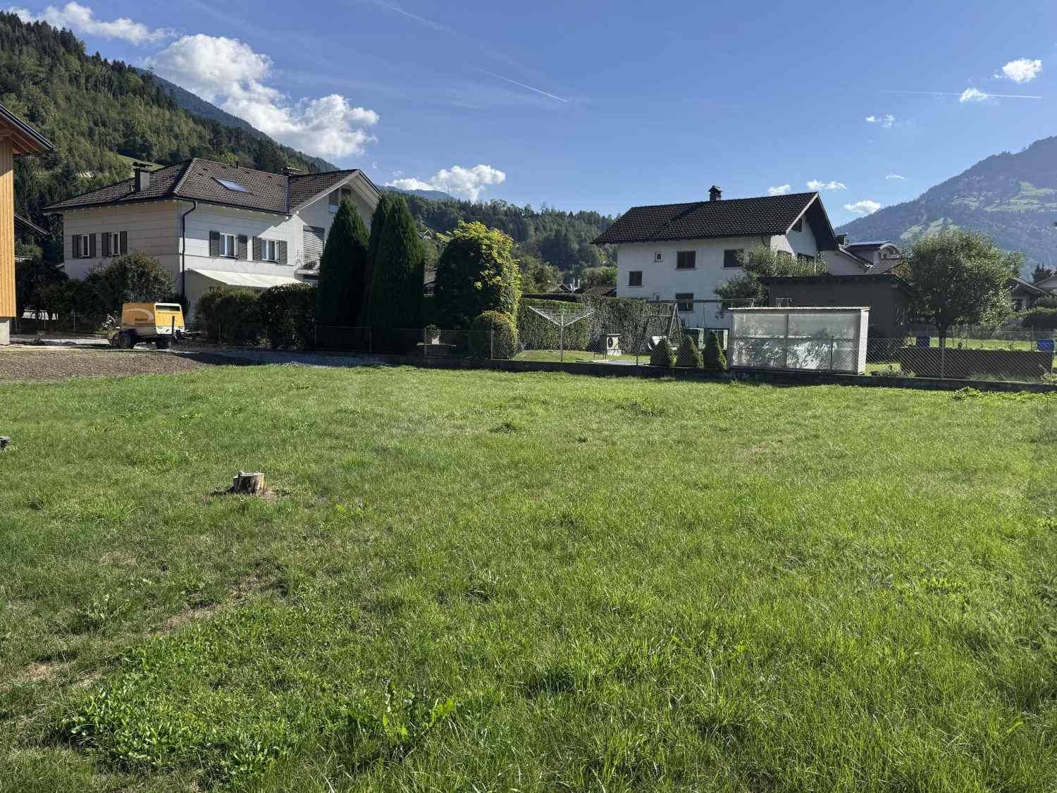 Weitläufige Rasenfläche mit Wohnhäusern, Bergen und einem kleinen Gewächshaus im Hintergrund.
