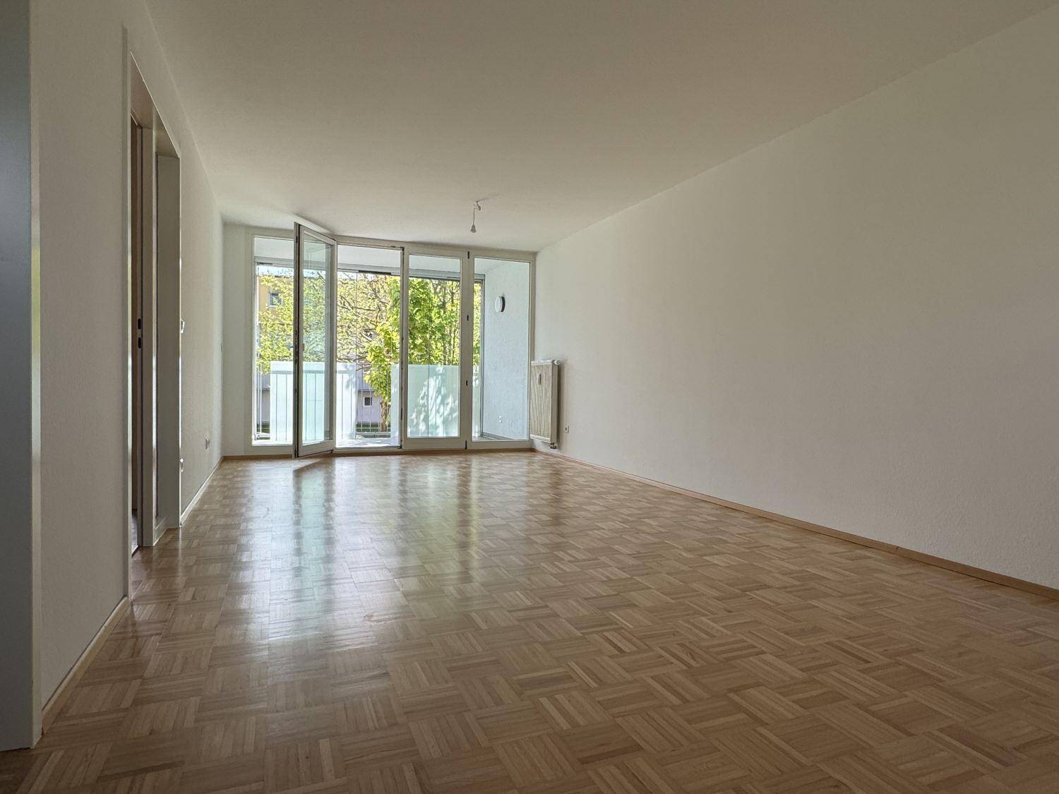 Geräumiges Wohnzimmer mit Parkettboden, weißen Wänden und Zugang zu einem Balkon.
