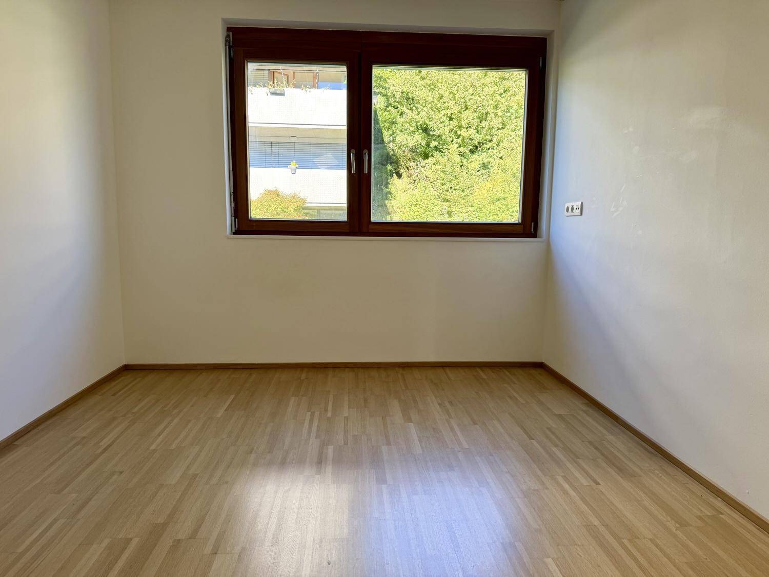 Leeres Zimmer mit Holzboden und Fenster, das einen grünen Ausblick auf Bäume bietet.