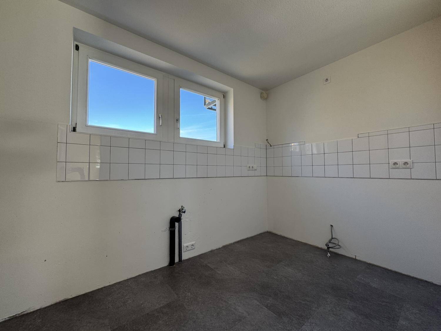 Leerer Küchenraum mit Fliesenwand, Anschlüssen für Geräte und einem großen Fenster.