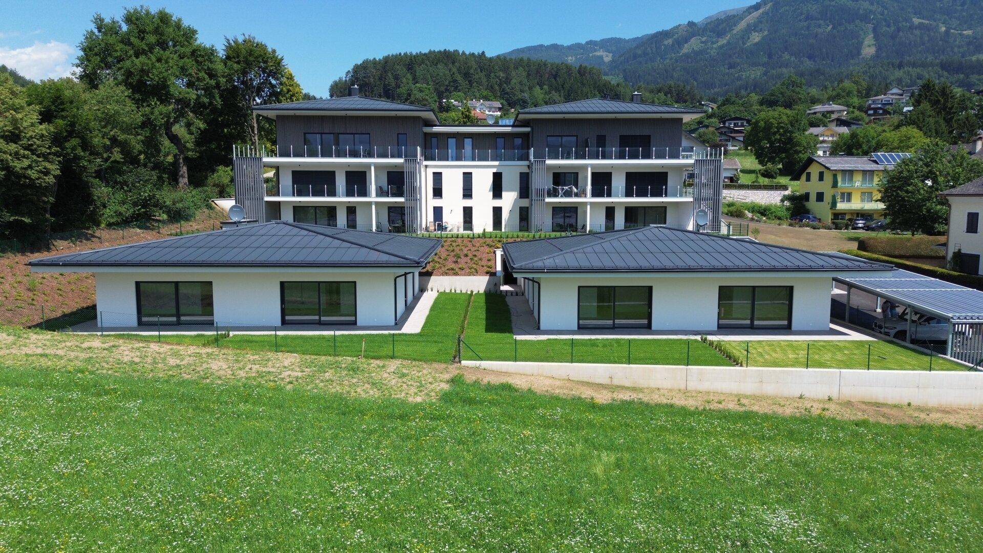 Moderne Wohnanlage mit zwei Mehrfamilienhäusern und zwei Bungalows in grüner Umgebung mit Bergblick.