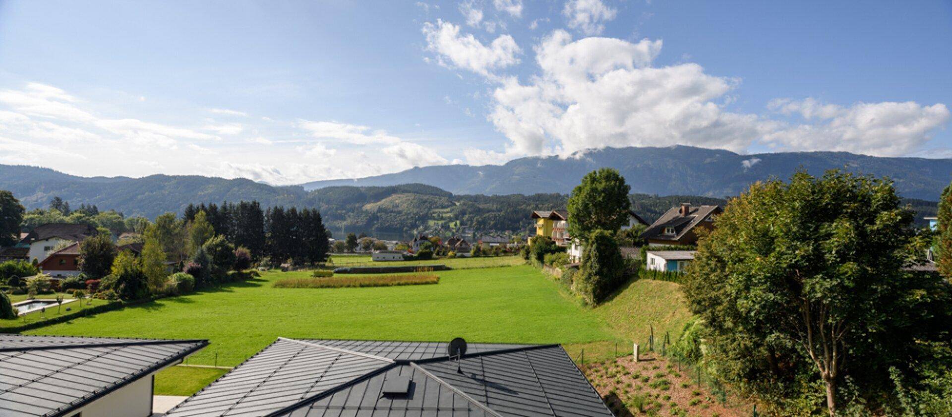 Weitläufiger Blick über grüne Felder und Dächer auf die umliegende Berglandschaft unter blauem Himmel.