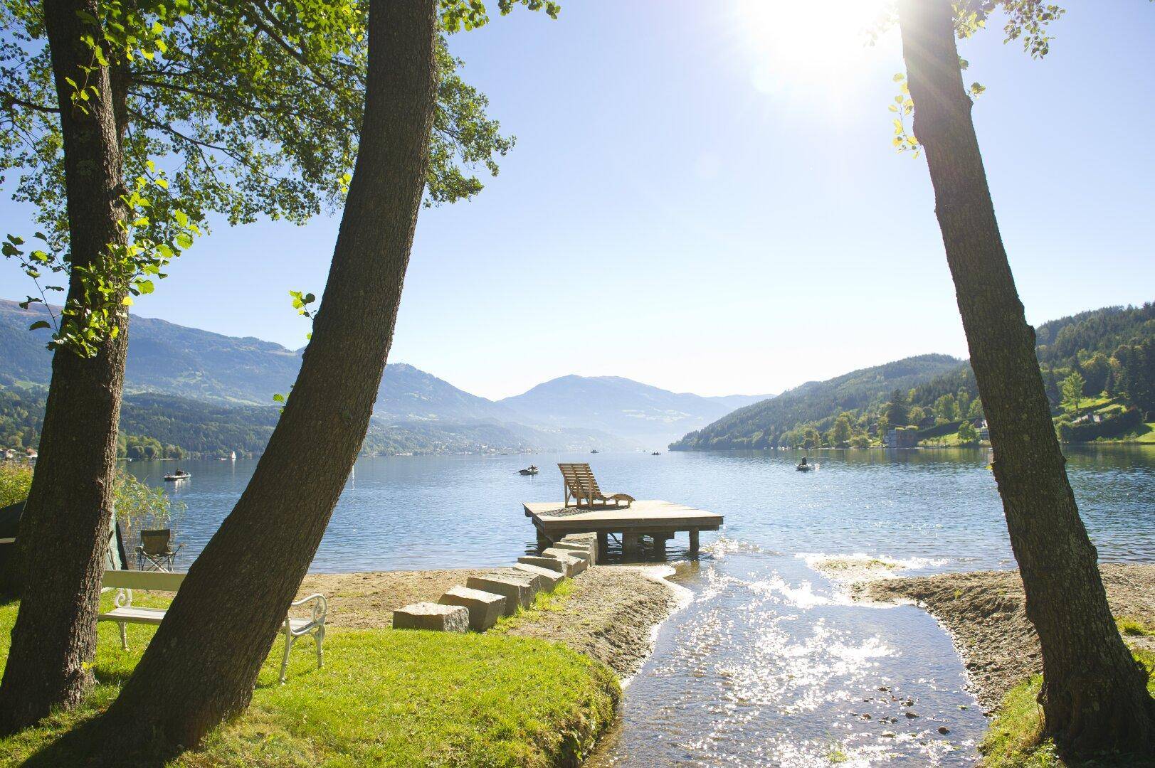 Idyllischer See mit kleinem Steg und Liegestuhl, umgeben von Bäumen und Bergen.