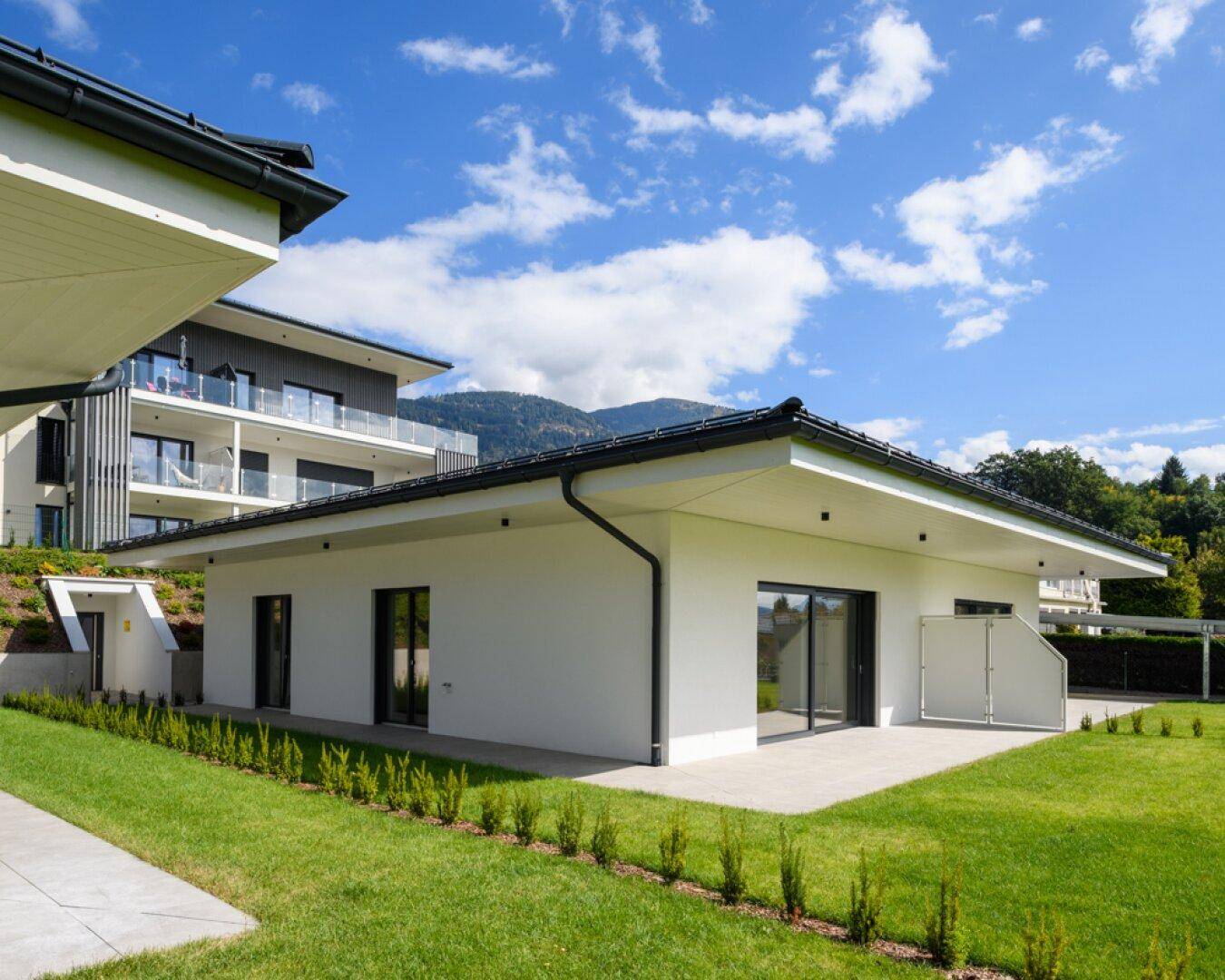 Moderne weiße Villa mit Flachdach, großen Glasfronten und gepflegtem Gartenbereich vor Bergkulisse.