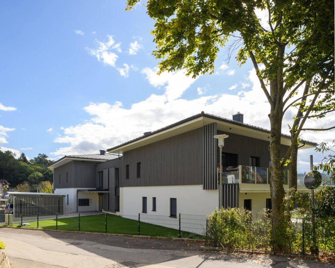 Seitenansicht eines modernen Gebäudes mit dunkler Holzfassade und Balkon, umgeben von Bäumen.