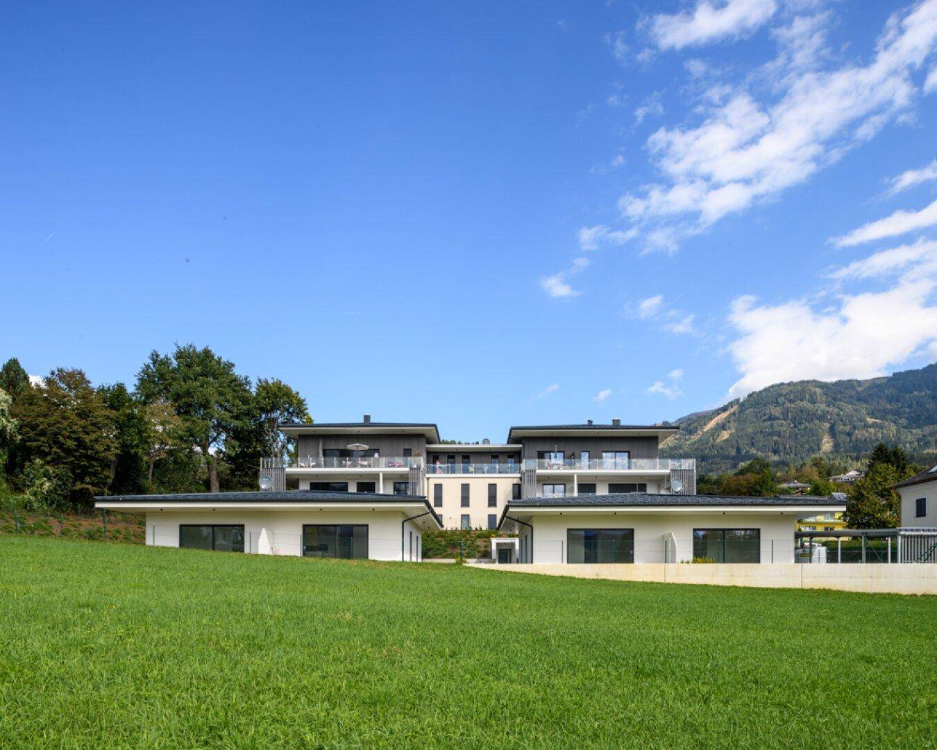 Moderner Wohnkomplex mit Bungalows und einem Apartmenthaus, umgeben von grünen Wiesen und Bergen.