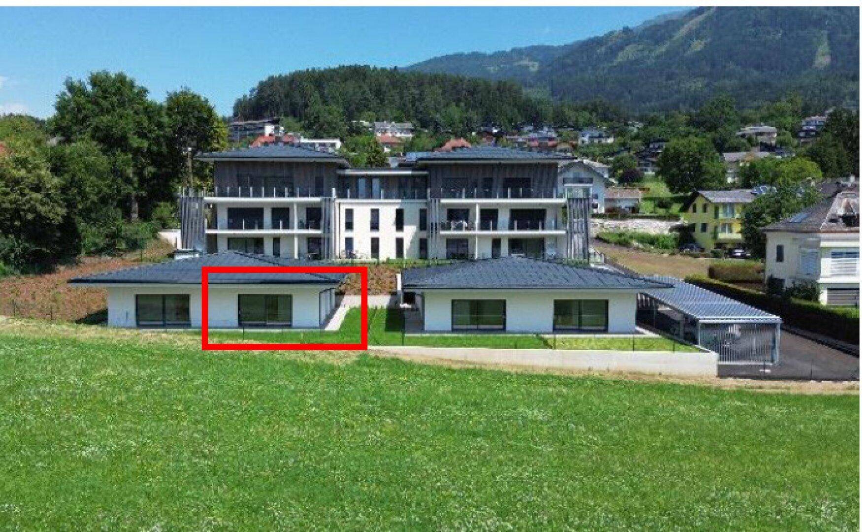 Luftaufnahme eines modernen Wohnkomplexes mit Bungalows und einem Mehrfamilienhaus vor einer Bergkulisse.