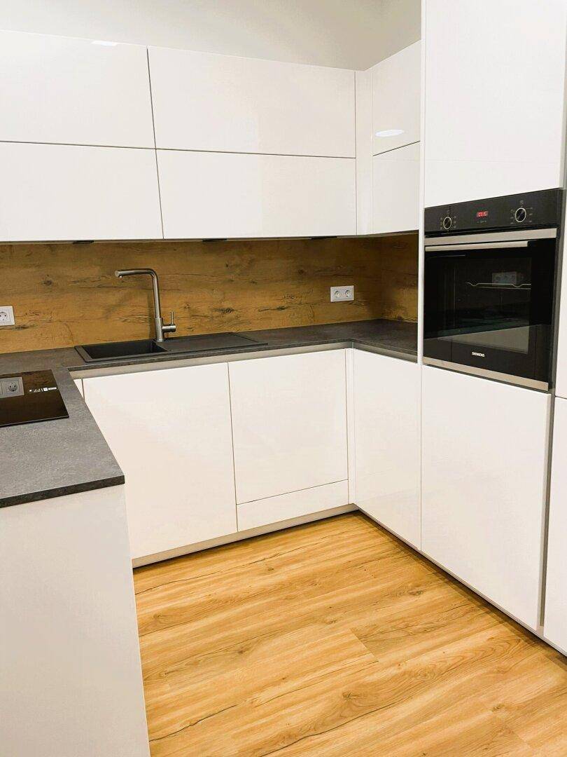 Moderne Küche mit weißen Hochglanzfronten, dunkler Arbeitsplatte und Holzakzenten an der Wand.
