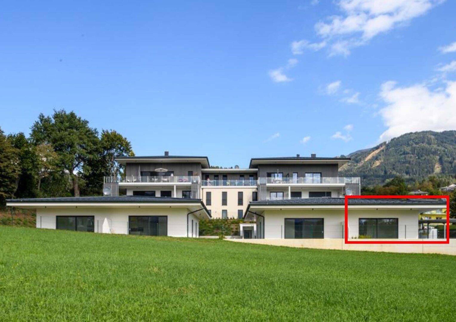 Moderne Wohnanlage mit weißer Fassade, umgeben von grünen Wiesen und Bergen im Hintergrund.