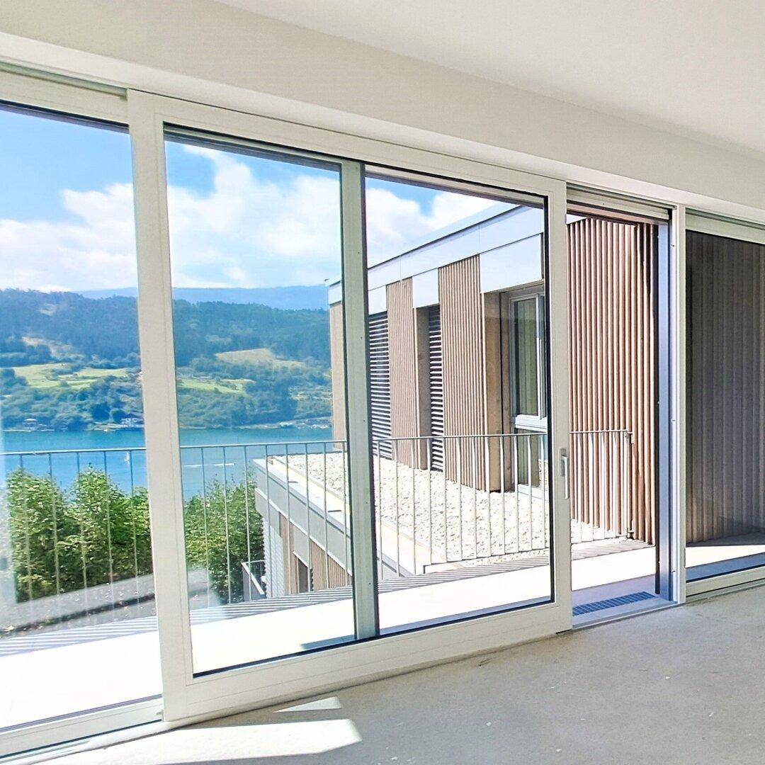 Heller Innenraum mit großen Glasfronten, die Zugang zu einem Balkon und Seeblick bieten.