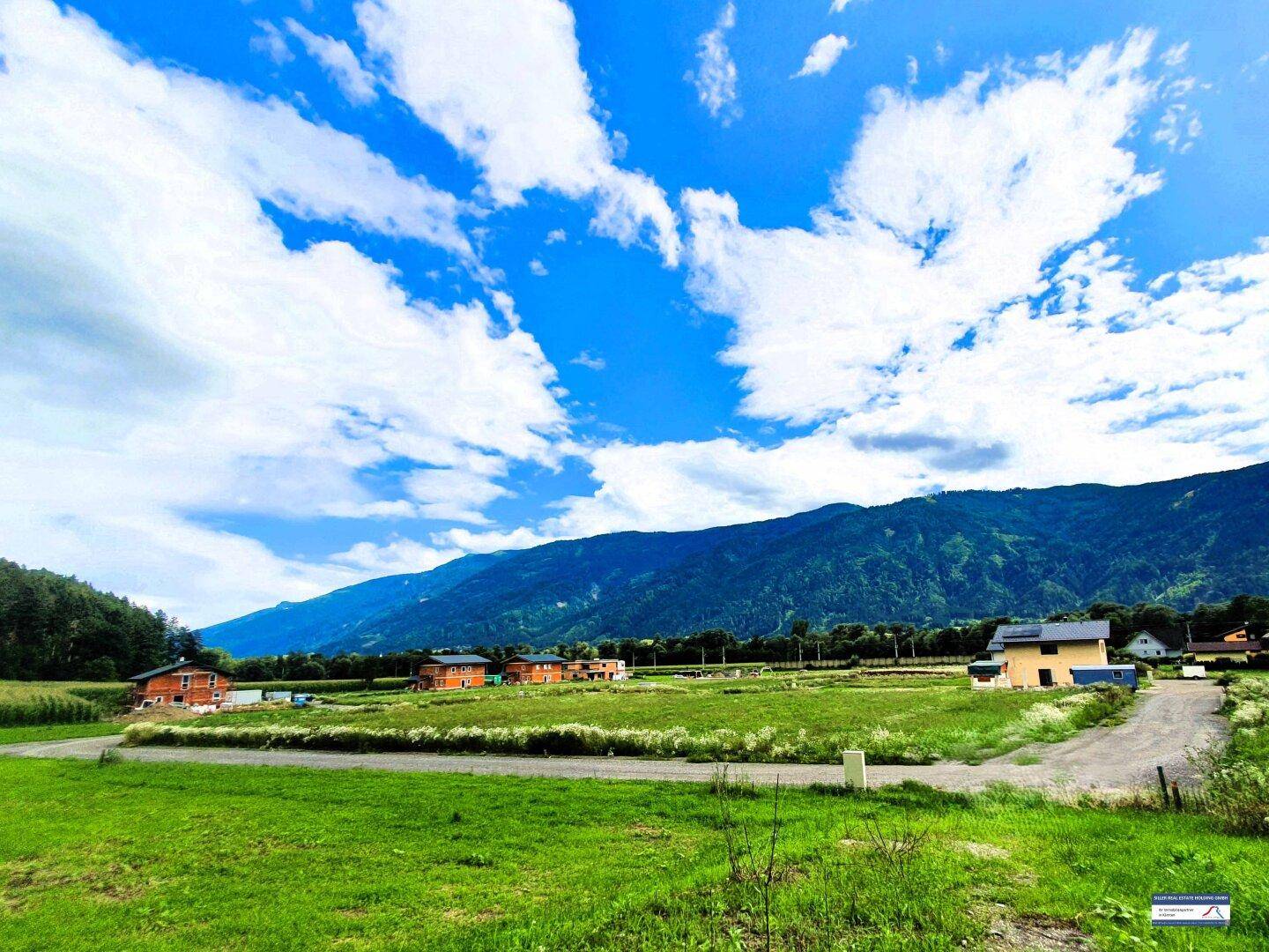 Weite Landschaft mit grünen Feldern, neuen Häusern und majestätischen Bergen unter strahlend blauem Himmel.