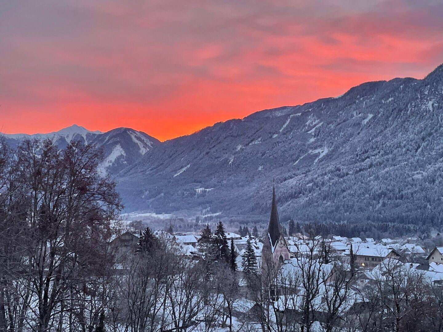Winterliche Dorflandschaft mit schneebedeckten Bergen unter einem dramatischen orange-roten Himmel bei Sonnenuntergang.