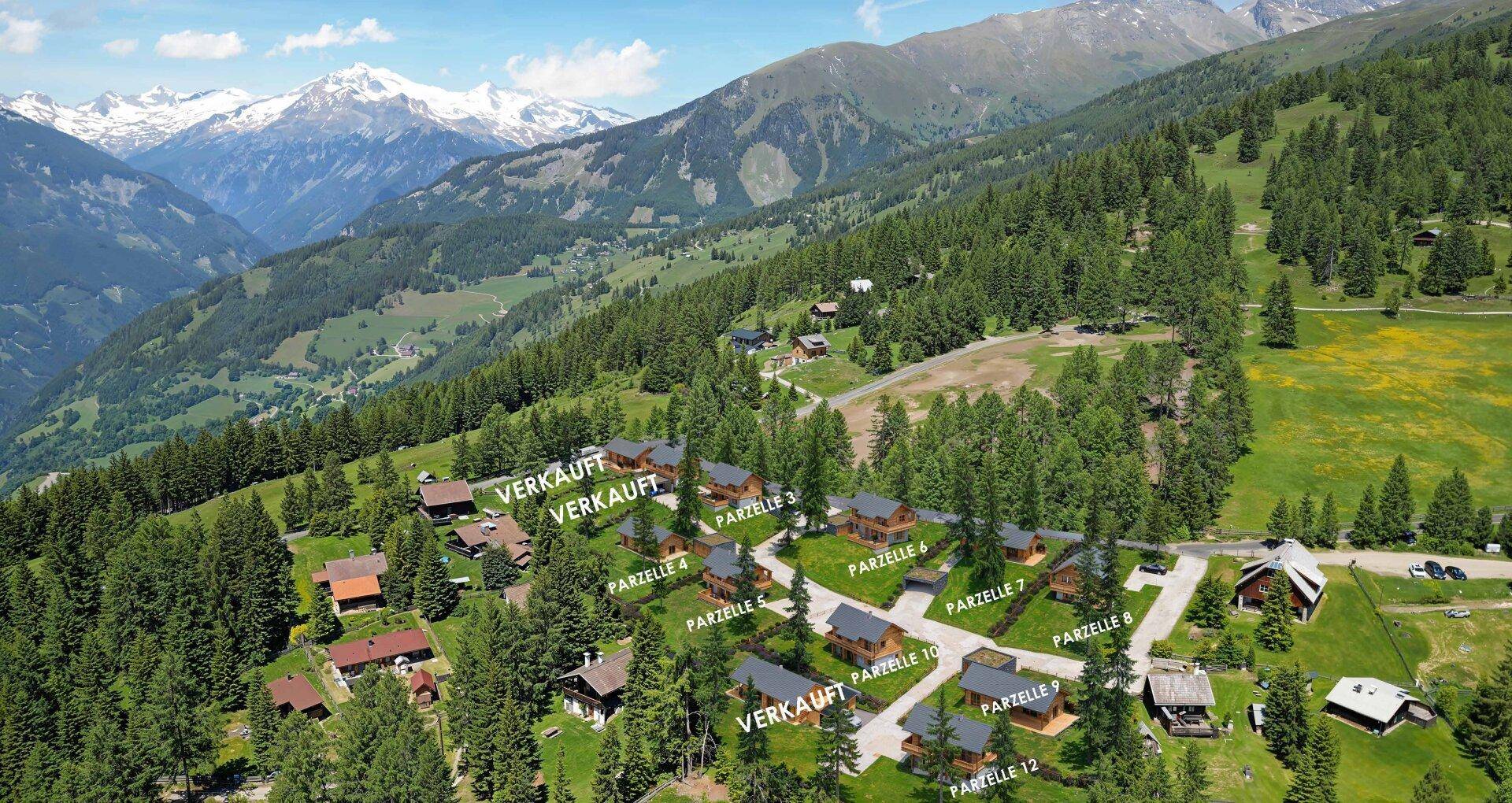 Luftaufnahme einer Siedlung von Holzhäusern in einer alpinen Berglandschaft mit schneebedeckten Gipfeln.