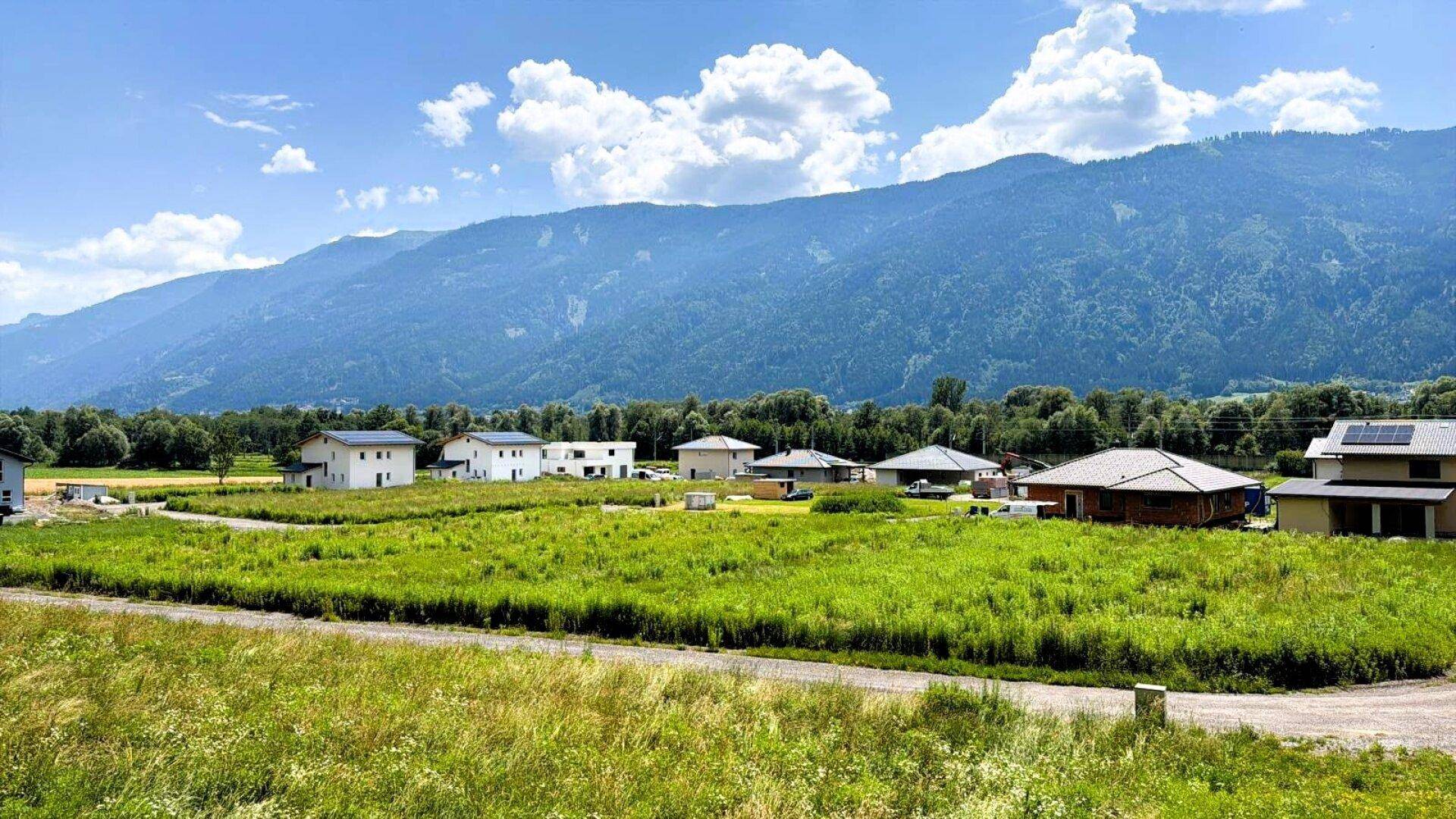Idyllische Landschaft mit Neubauten und weiten Wiesenflächen, umgeben von einer malerischen Berglandschaft.