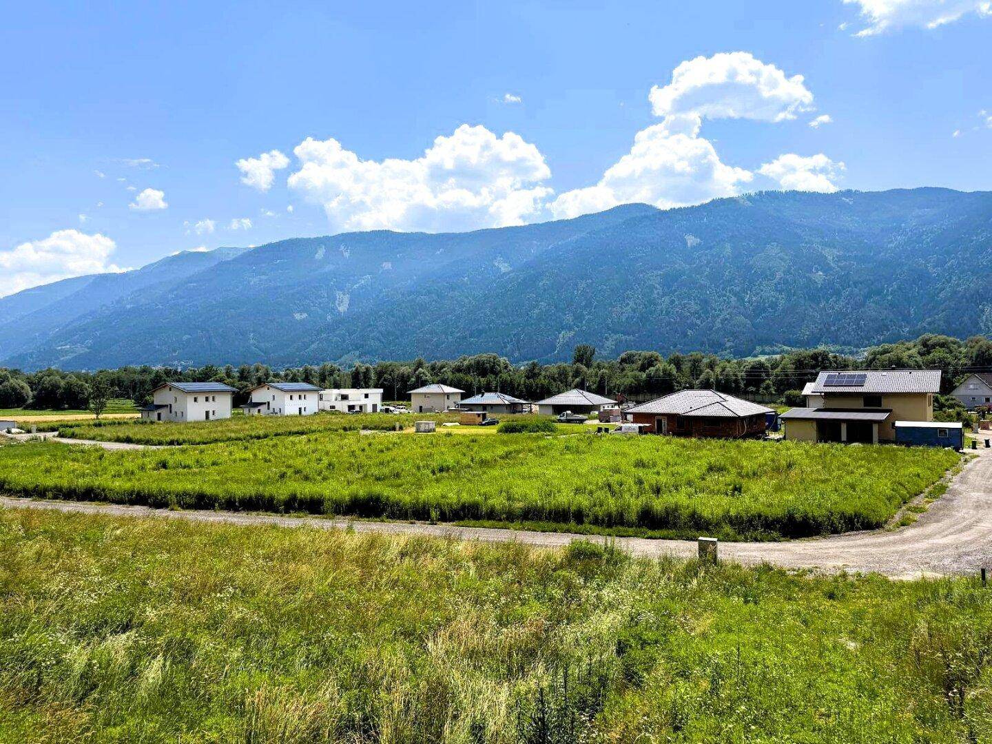 Weitläufige Landschaft mit grünen Wiesen, modernen Häusern und majestätischen Bergen im Hintergrund unter blauem Himmel.