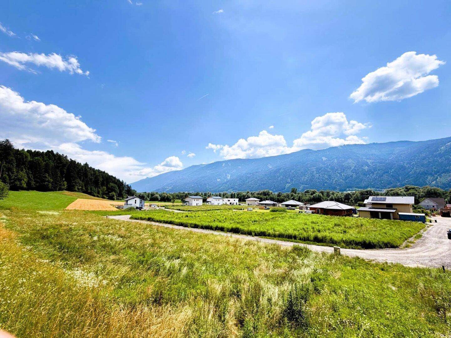Panoramablick auf eine ländliche Gegend mit Neubauten, grünen Feldern und bewaldeten Bergen unter blauem Himmel.
